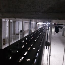 Instak - Metalowa korytka kablowa zamocowana pod stropem, widok z dołu, oświetlenie budowlane, elementy konstrukcyjne w tle. Perspektywa zbieżna, ciemna tonacja.