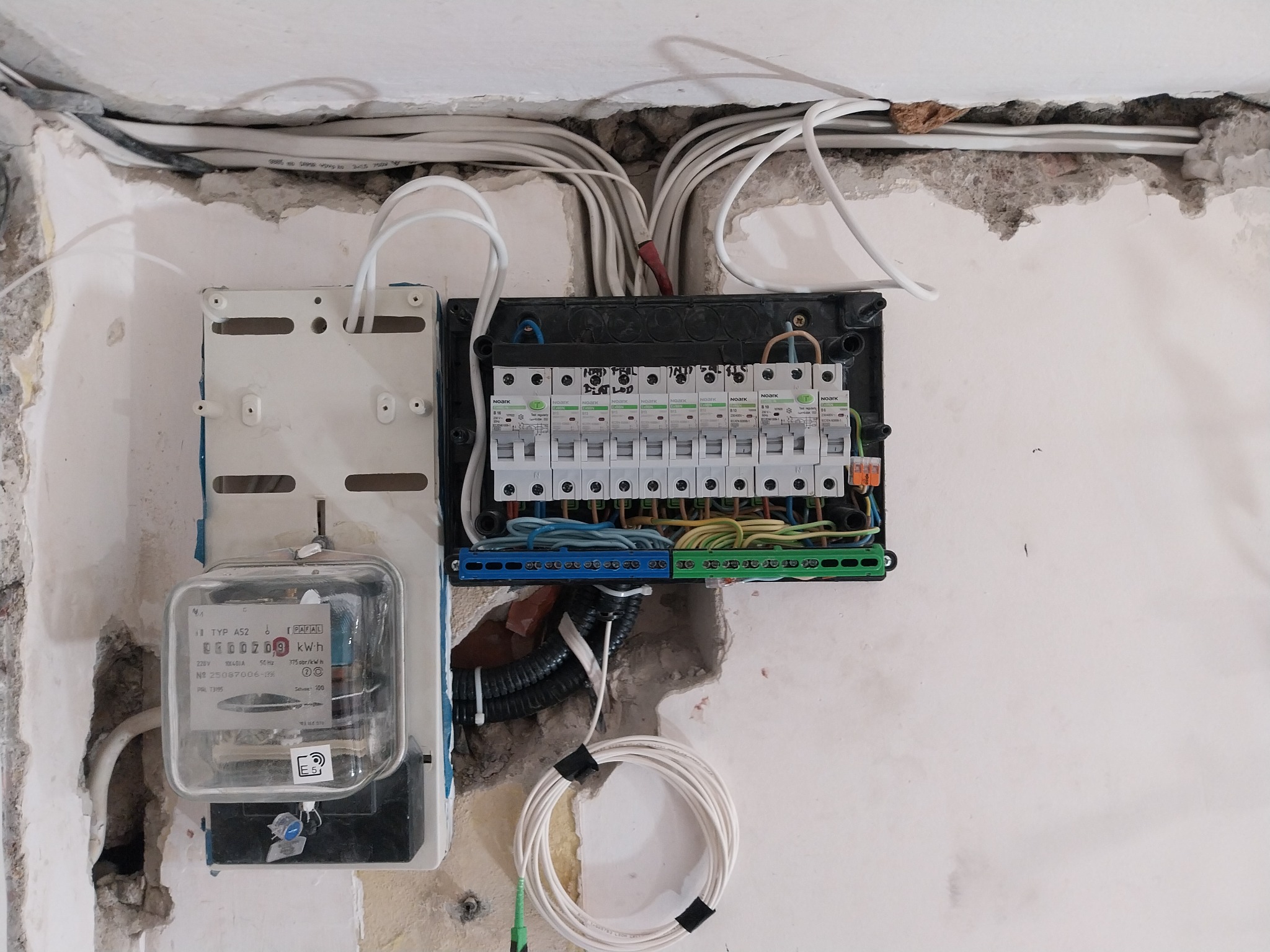 Rozdzielnia elektryczna z licznikiem na ścianie w trakcie montażu. Widoczne przewody, bezpieczniki i elementy instalacji. Ściana z widocznymi śladami prac remontowych.