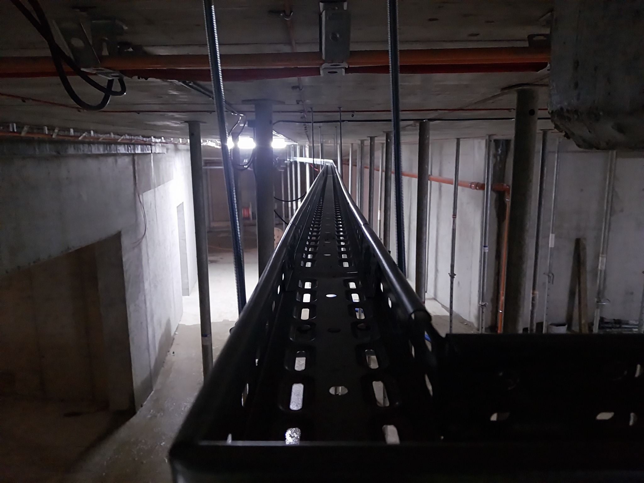 Metalowa korytka kablowa zamontowana pod sufitem w surowym wnętrzu budynku w trakcie budowy. Widoczne wsporniki i instalacje pod stropem.
