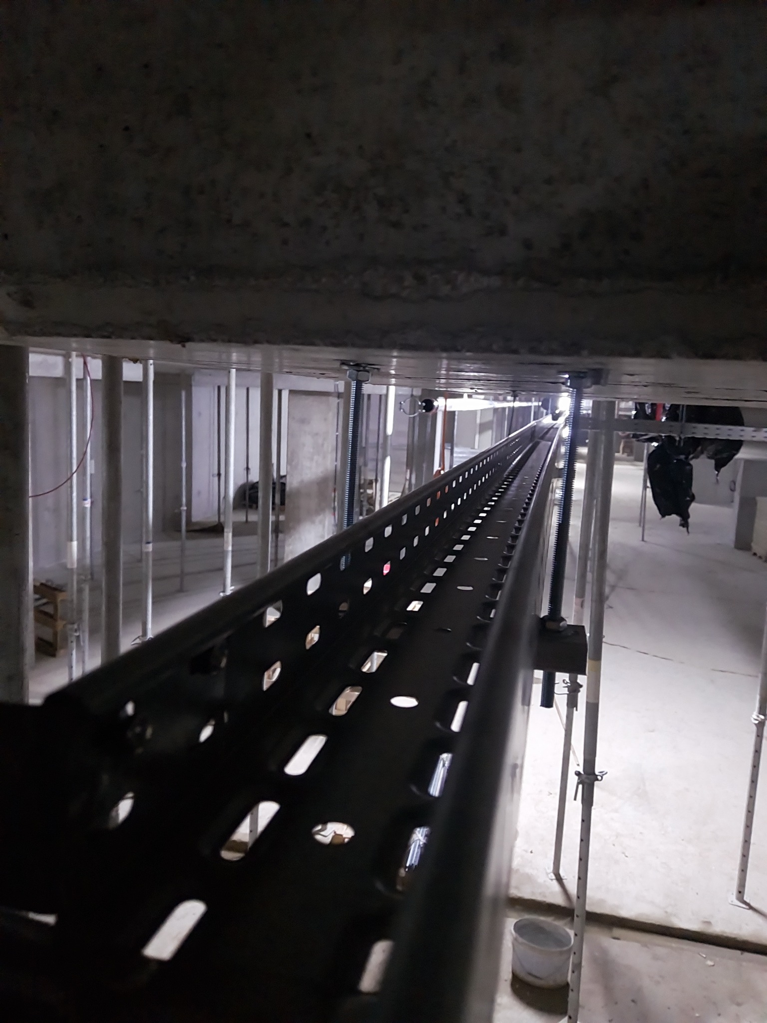 Metalowa korytka kablowa zamocowana pod stropem, widok z dołu, oświetlenie budowlane, elementy konstrukcyjne w tle. Perspektywa zbieżna, ciemna tonacja.