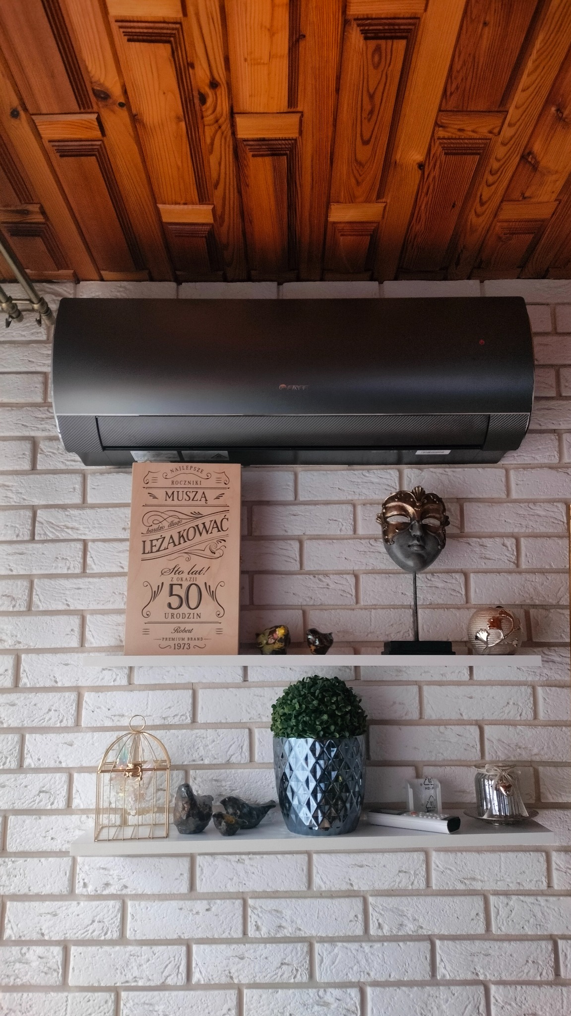 Czarny klimatyzator Gree na białej ceglanej ścianie z drewnianym sufitem. Dekoracyjne półki z ozdobami dodają wnętrzu charakteru.