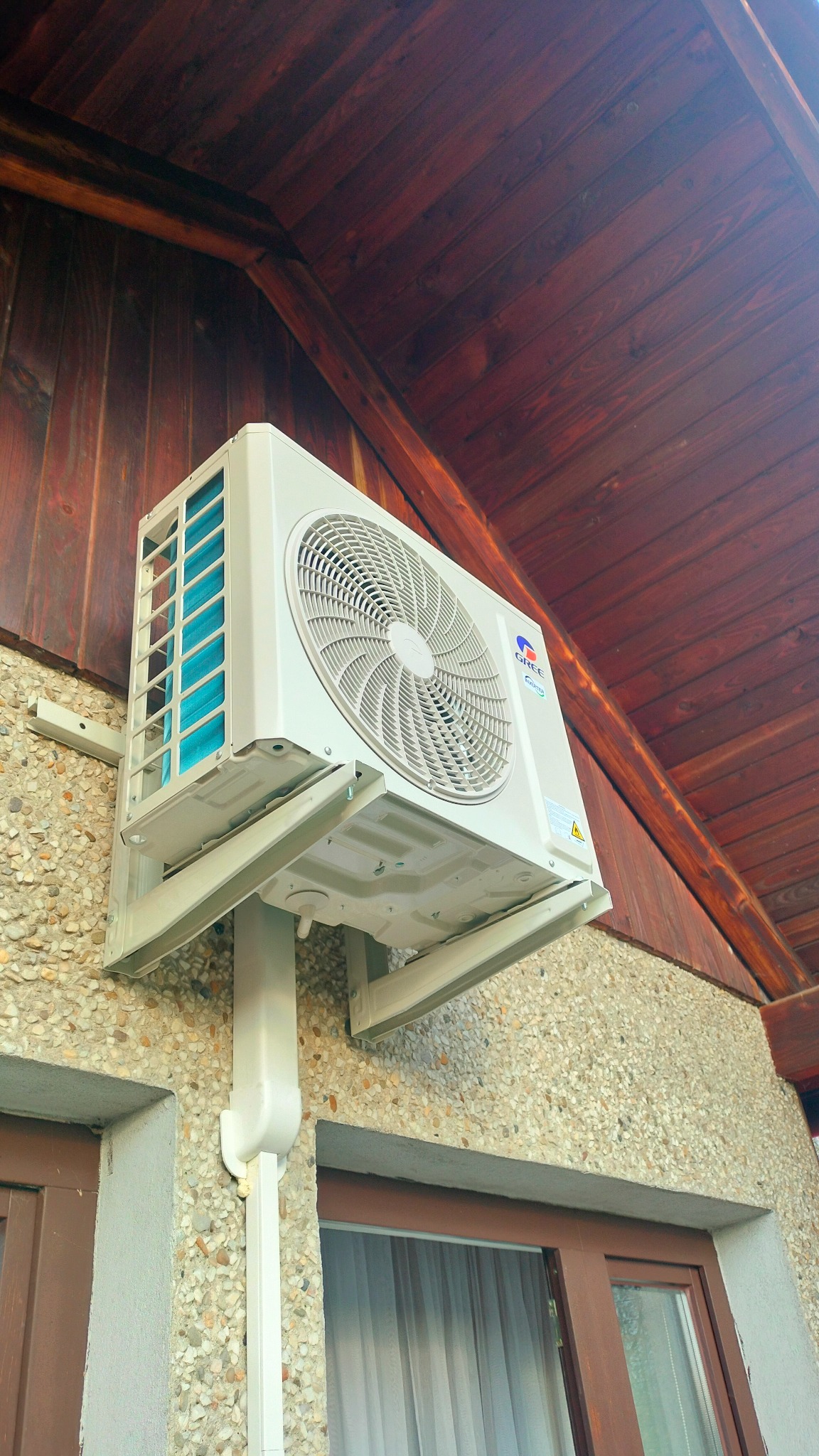 Biały klimatyzator Gree zamontowany na elewacji budynku z oknami i drewnianym podbitką dachu, widok z dołu.