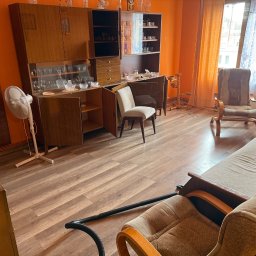 Sheremet Transport - Pokój z meblami w stylu retro przed opróżnianiem. Widoczny segment, krzesła, sofa i rura od odkurzacza. Pomarańczowe ściany i drewniana podłoga. Częściowy widok.