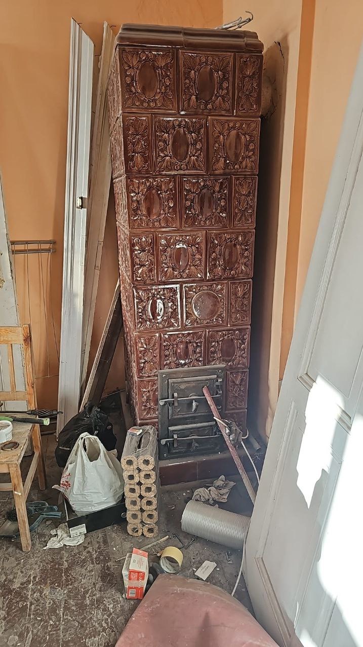 Brązowy, kaflowy piec w trakcie demontażu/przygotowania do transportu. Wokół porozrzucane przedmioty, elementy wyposażenia wnętrza i materiały budowlane.