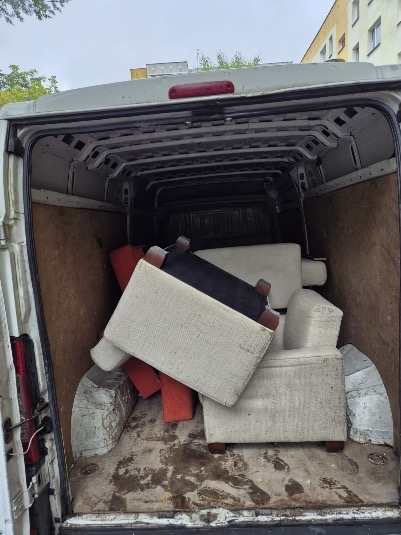 Wnętrze brudnego busa z meblami: fotel, pufy w beżowej i czarnej tapicerce. Widoczne ślady użytkowania na podłodze i ścianach pojazdu. Ujęcie od tyłu, otwarte drzwi.