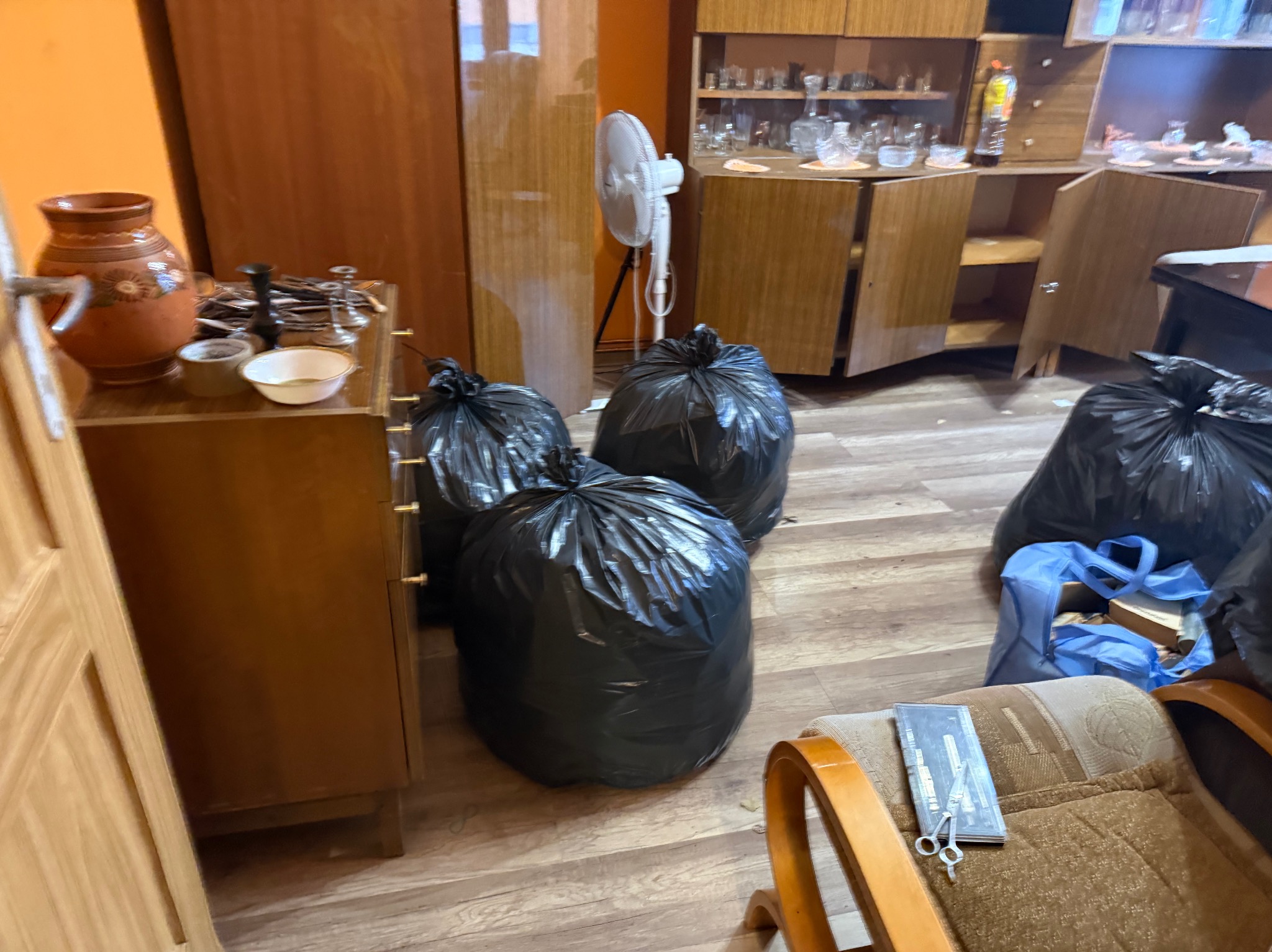 Pokój przed opróżnianiem: meble, bibeloty i worki na śmieci. Na krześle leżą nożyczki i opakowanie. Widoczny wentylator i otwarte szafki.