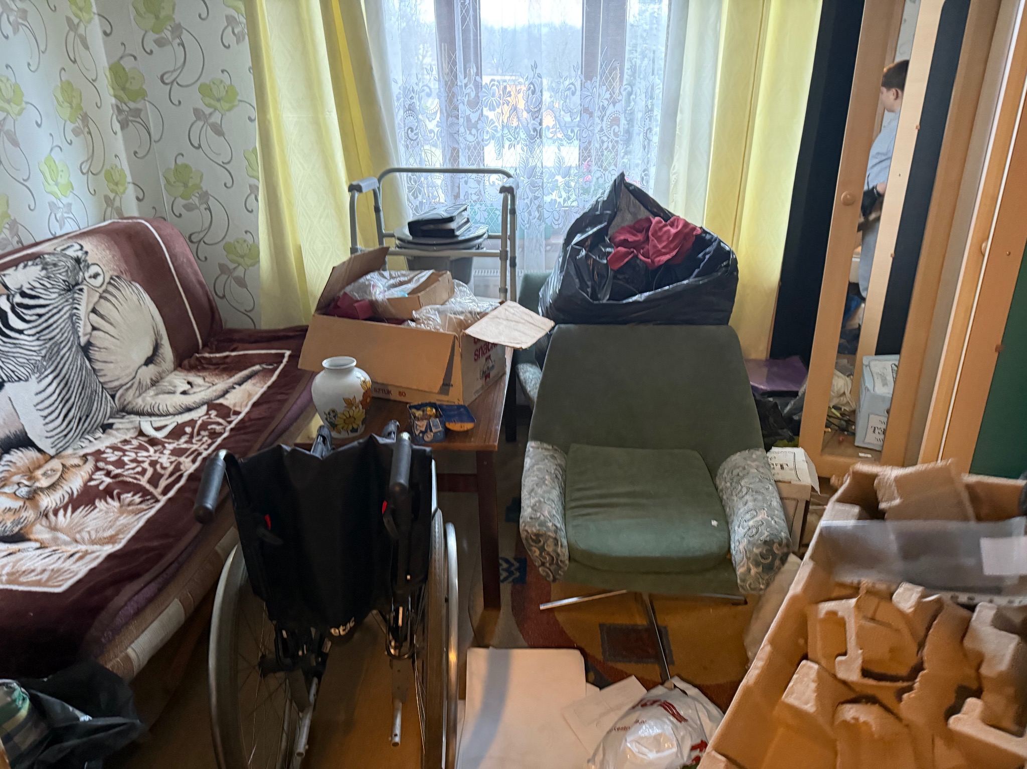 Pokój przed opróżnianiem: fotel, sofa, wózek inwalidzki, kartony i worki ze śmieciami. Widoczne okno z firanką i wzorzysta tapeta. Przygotowanie do opróżniania mieszkania.