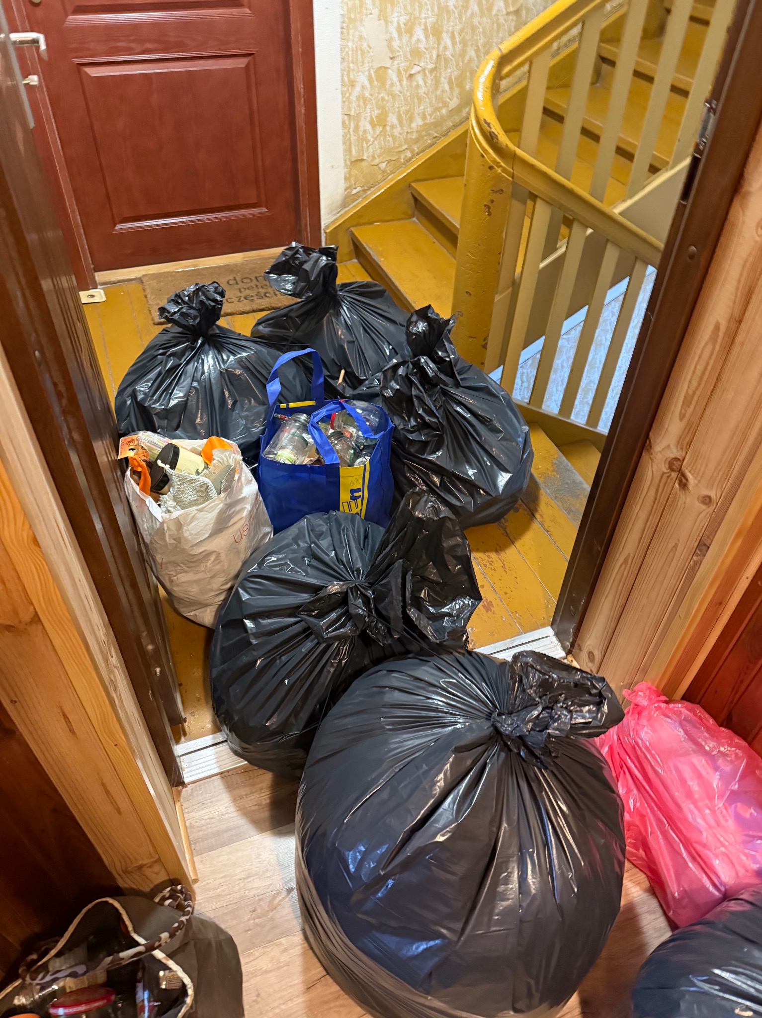 Wnętrze klatki schodowej z workami śmieci, torbami i przedmiotami do usunięcia. Widoczne schody i drzwi. Perspektywa z góry, ukazująca etap opróżniania mieszkania.