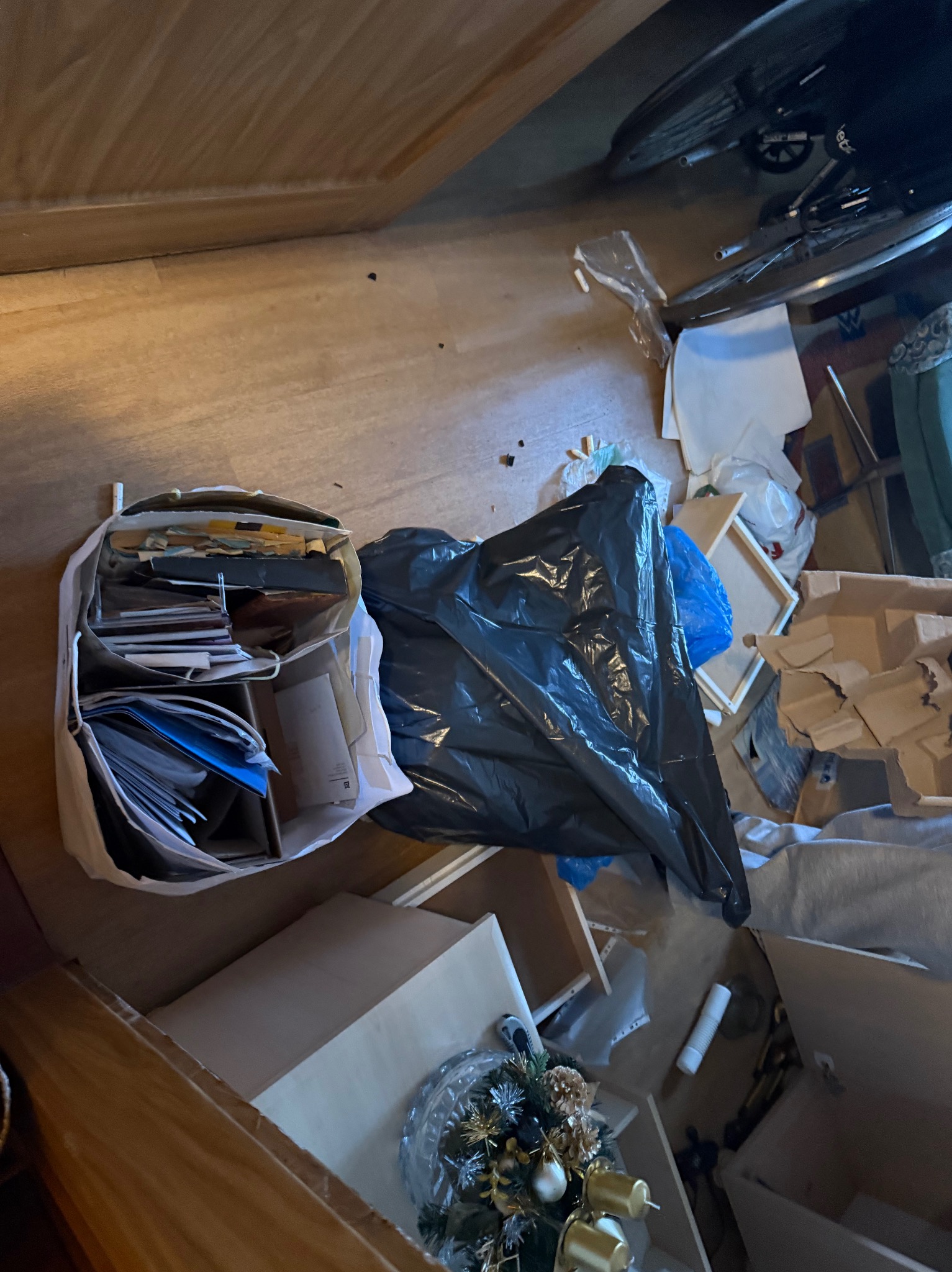 Opróżniane mieszkanie: torba z dokumentami, czarny worek na śmieci, kartony i dekoracja świąteczna na podłodze. Widok z góry na bałagan w trakcie opróżniania.