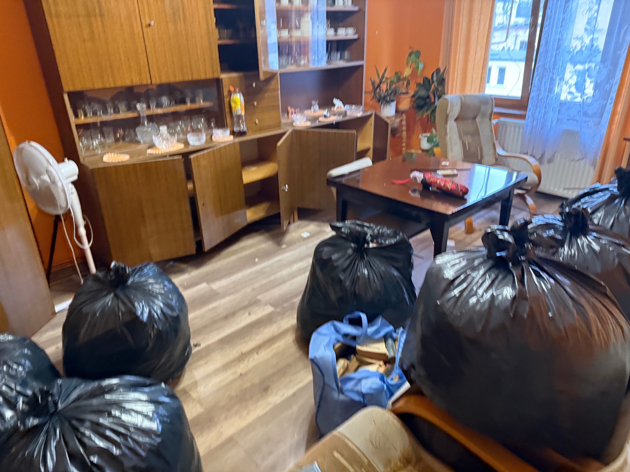 Pokój przed opróżnianiem: meblościanka z wyposażeniem, stół, krzesło i worki ze śmieciami. Widoczny biały wentylator i okno z firanką. Pomieszczenie w trakcie porządkowania.