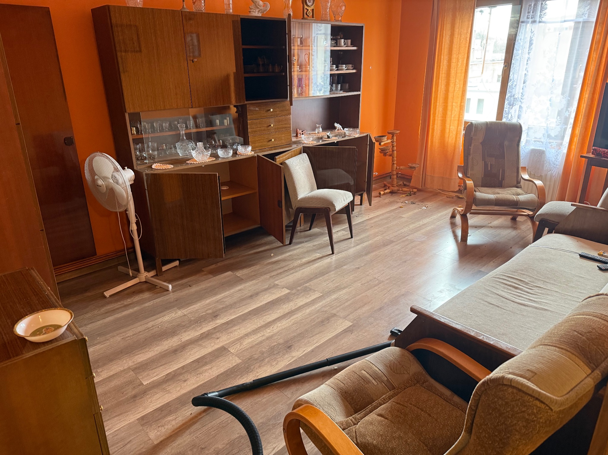 Pokój z meblami w stylu retro przed opróżnianiem. Widoczny segment, krzesła, sofa i rura od odkurzacza. Pomarańczowe ściany i drewniana podłoga. Częściowy widok.