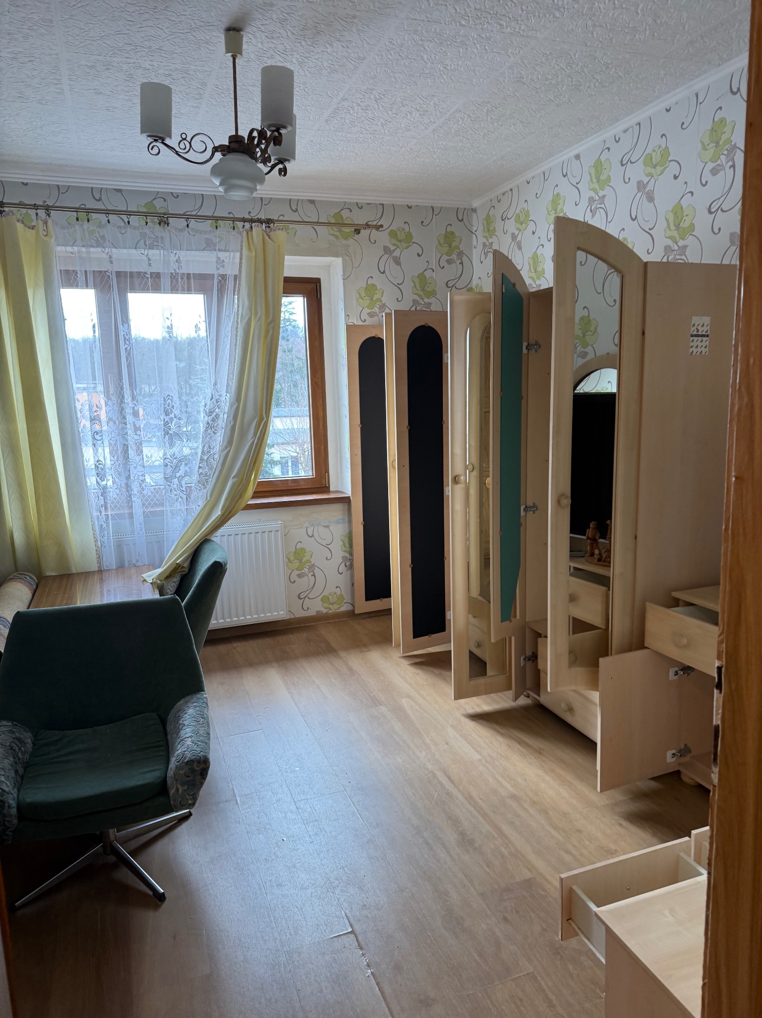 Pokój z rozłożonymi, jasnymi meblami do przechowywania, fotel z zieloną tapicerką, okno z firanką i zasłoną. Widoczne ślady użytkowania na podłodze.