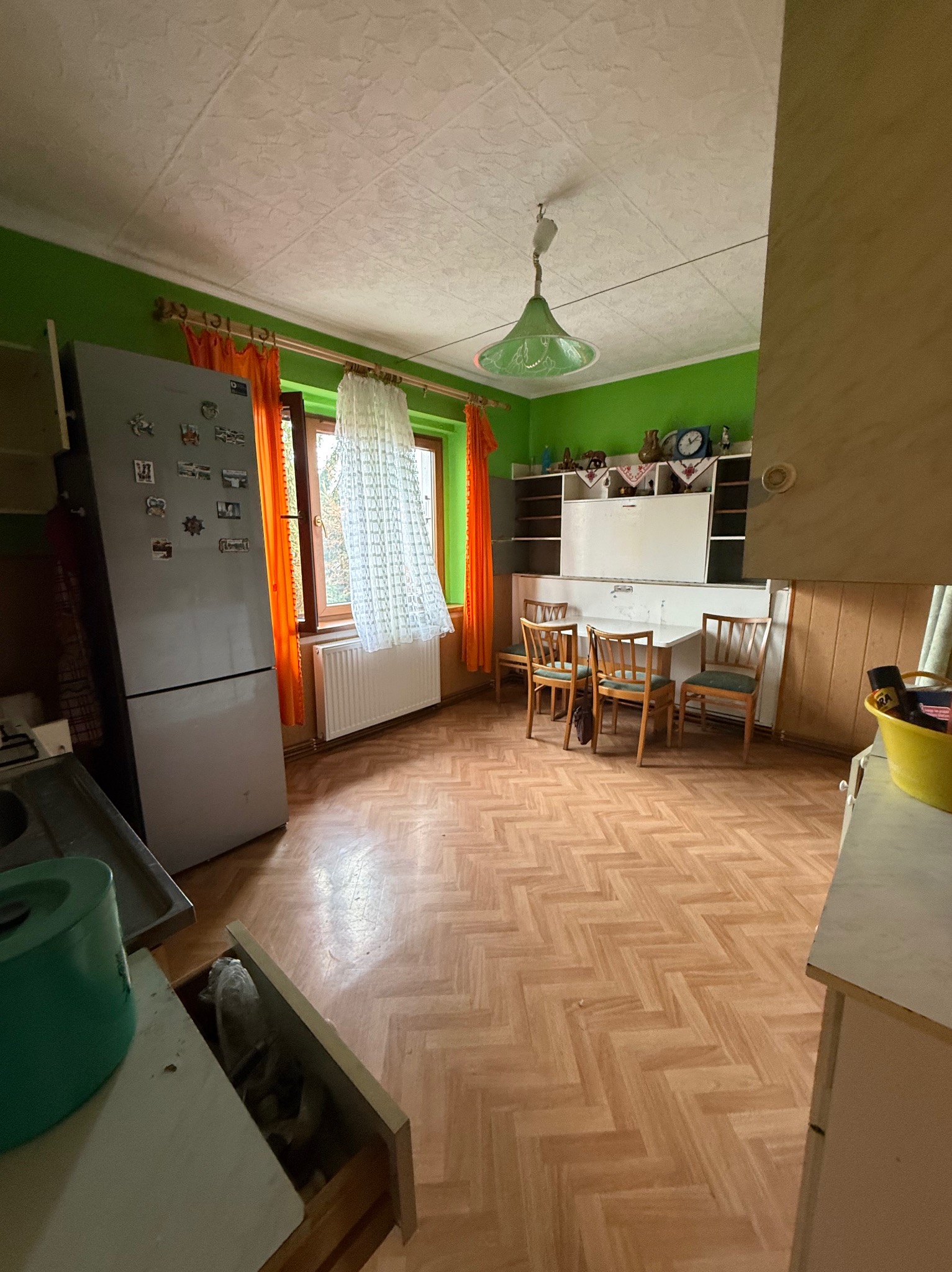 Wnętrze kuchni z zielonymi ścianami, drewnianą podłogą w jodełkę i pomarańczowymi zasłonami. Widoczny stół z krzesłami, lodówka i otwarte szuflady.