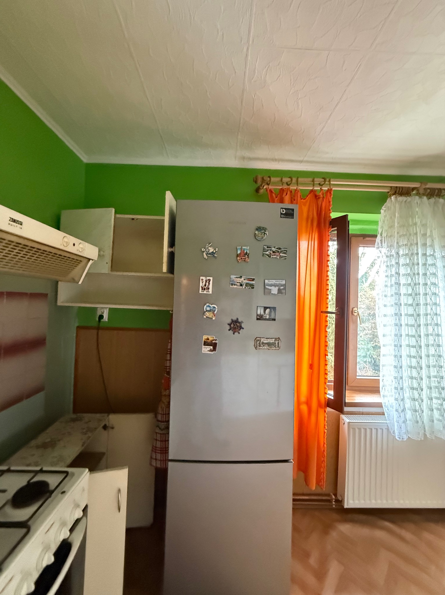 Kuchnia z zielonymi ścianami, lodówką z magnesami, oknem z pomarańczową i białą zasłoną. Widoczny fragment kuchenki i szafek. Wnętrze przed remontem.