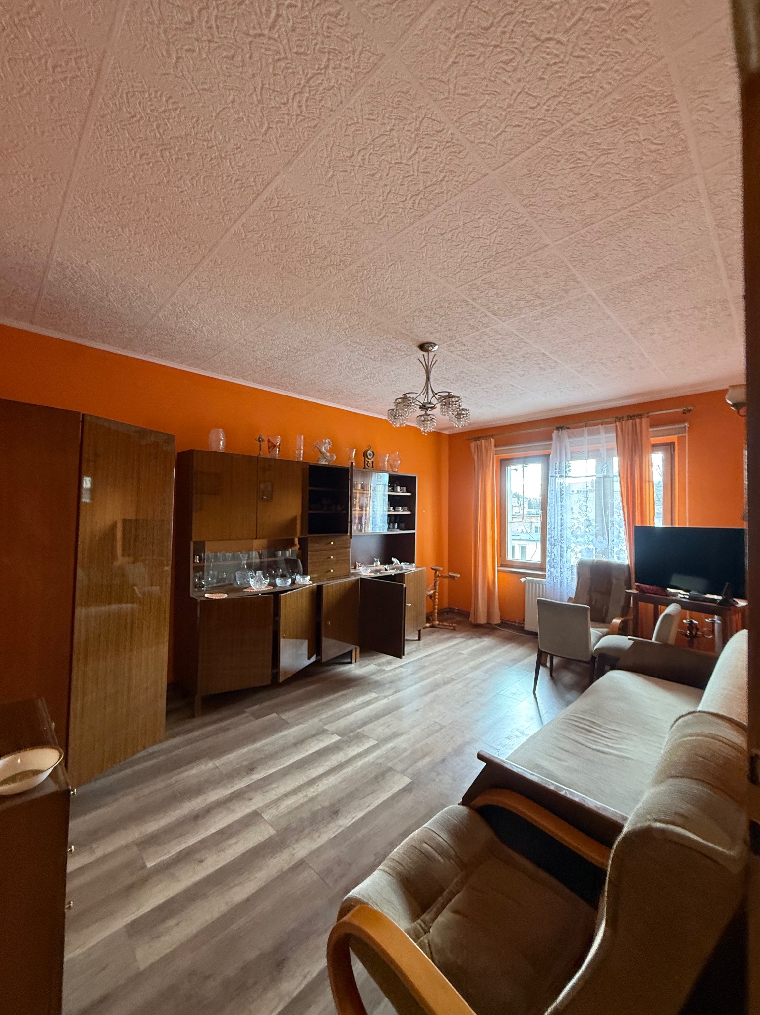 Umeblowane wnętrze pokoju z pomarańczowymi ścianami, drewnianą podłogą i meblami w stylu vintage. Widoczna sofa, szafa i okno z firankami.