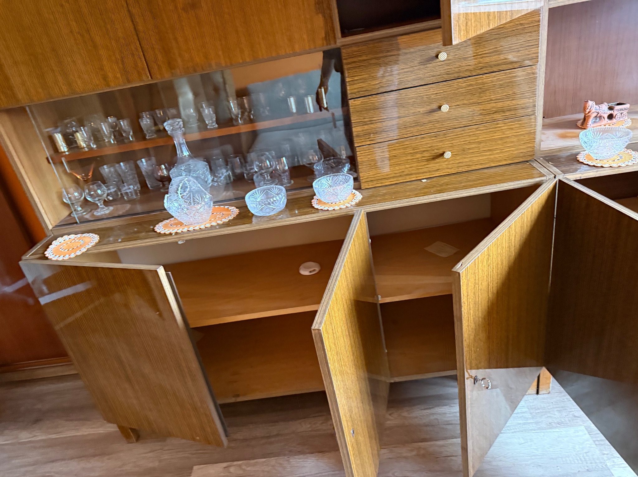 Staromodny, drewniany segment meblowy z otwartymi drzwiczkami i szklaną witryną wypełnioną szkłem dekoracyjnym. Widoczne puste półki i szuflady.