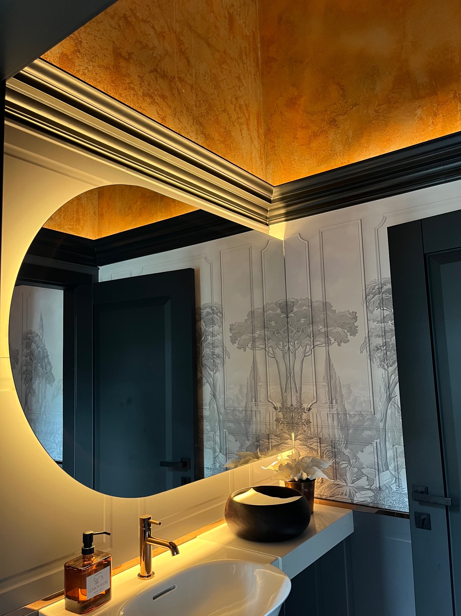 Stylowa łazienka z okrągłym lustrem podświetlanym LED, umywalką, złotą armaturą i dekoracyjną tapetą w odcieniach szarości. Sufit w kolorze miedzianym.