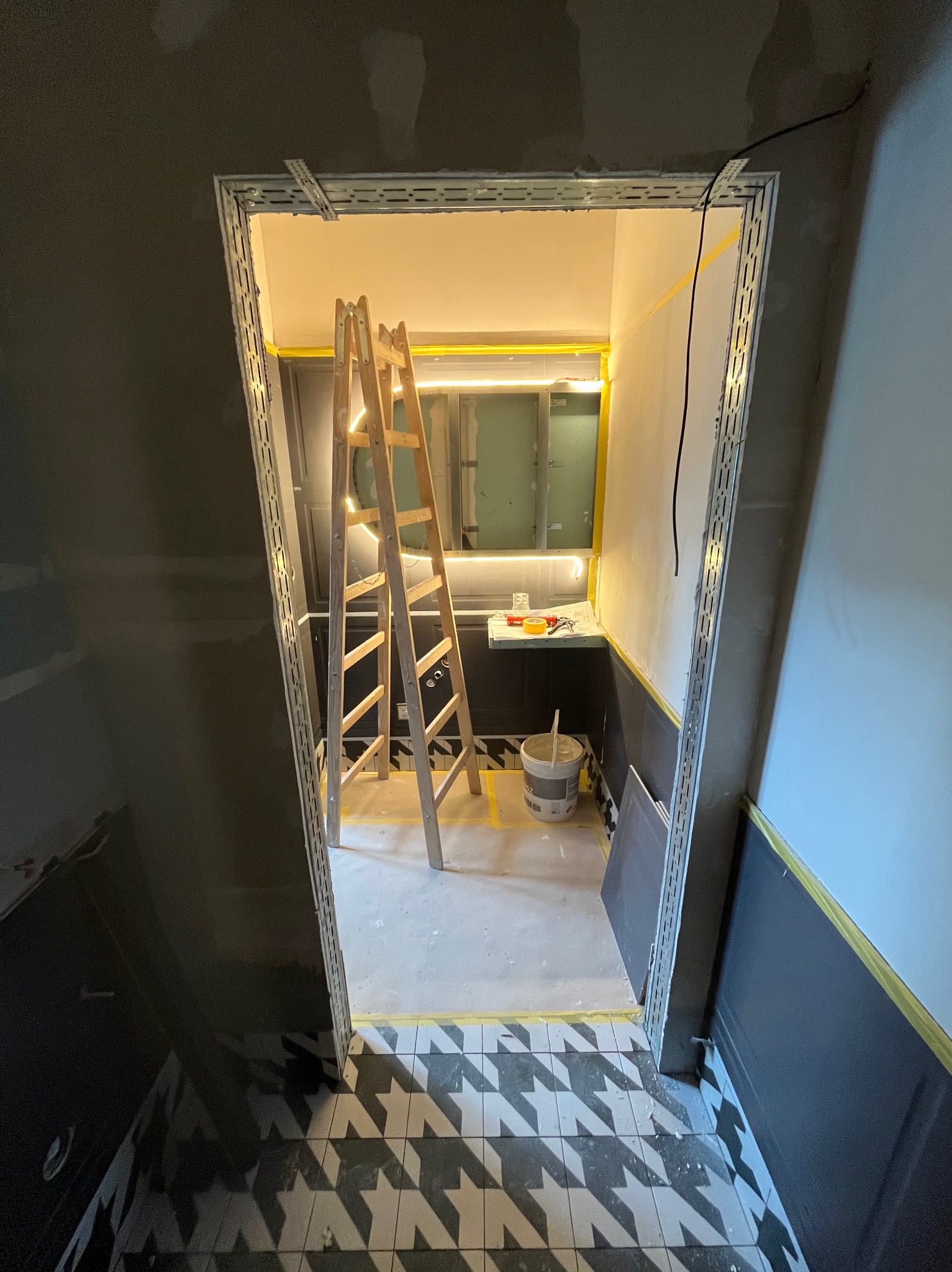 Łazienka w trakcie prac wykończeniowych: drabina, wiadro z zaprawą, płytki podłogowe w geometryczny wzór, oświetlenie LED. Widoczne profile montażowe.