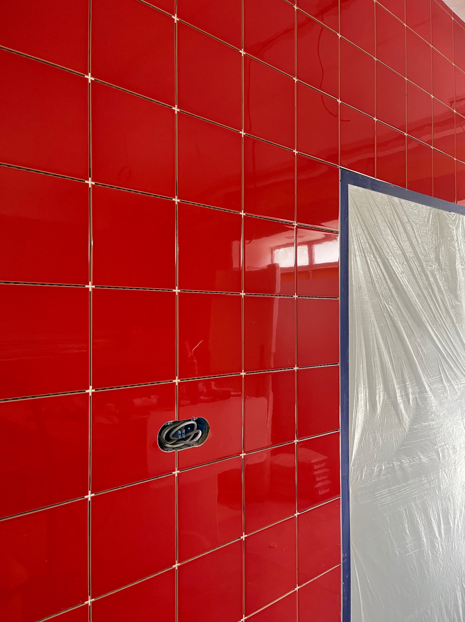 Ściana wyłożona błyszczącymi, czerwonymi płytkami z krzyżykami dystansowymi, widoczne gniazdko elektryczne i zabezpieczona folią malarską ściana.