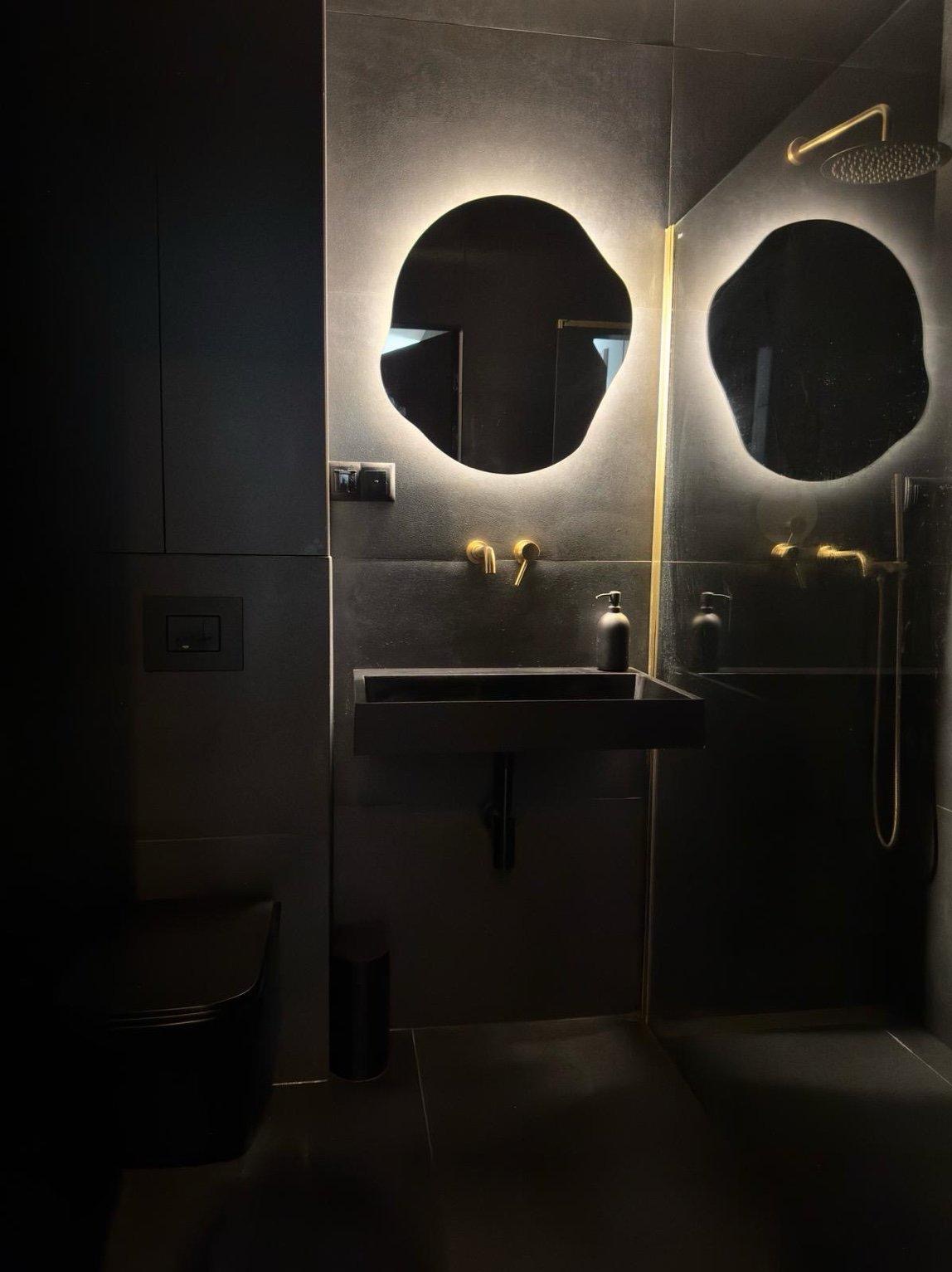 Nowoczesna, ciemna łazienka z podświetlanymi, nieregularnymi lustrami, czarną umywalką i złotą armaturą. Minimalistyczny design z akcentami luksusu.