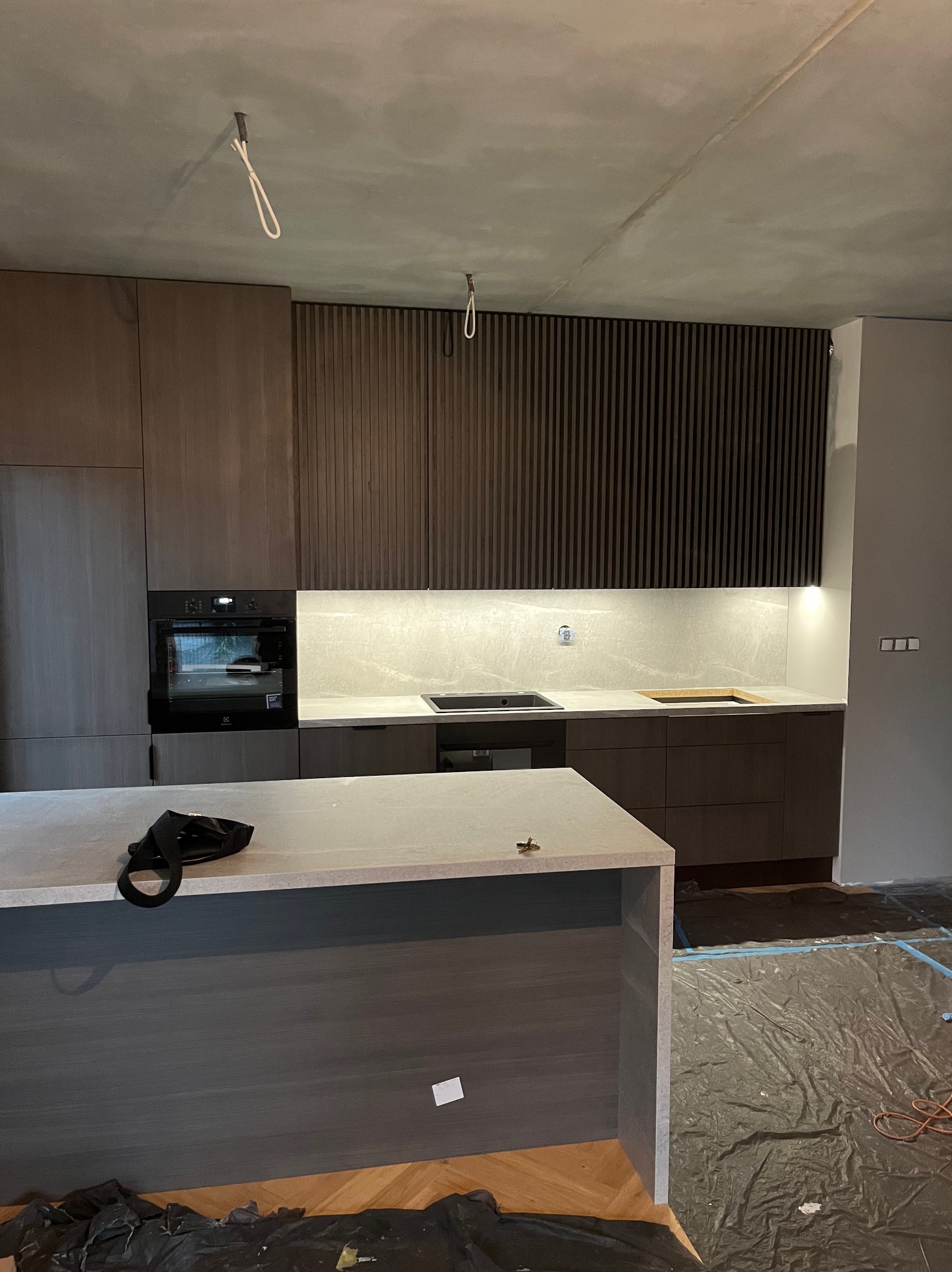 Nowoczesna kuchnia w trakcie montażu: ciemne, pionowe panele dekoracyjne, jasny blat, wyspa kuchenna. Podłoga zabezpieczona folią malarską, widoczne kable oświetleniowe.