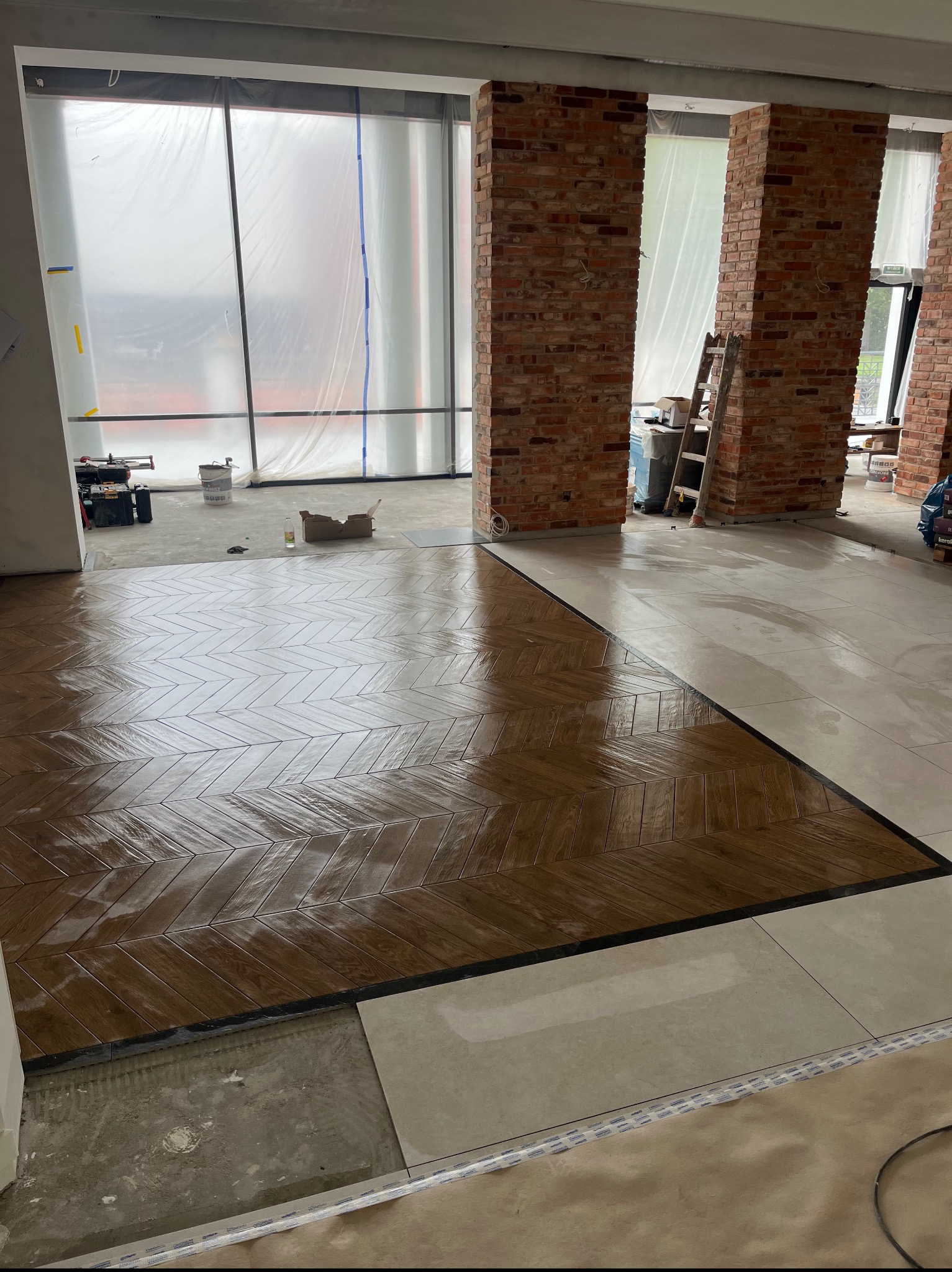 Układanie parkietu w jodełkę i płytek w przestronnym wnętrzu z ceglanymi filarami. Okna zabezpieczone folią malarską. Częściowo ułożona podłoga.
