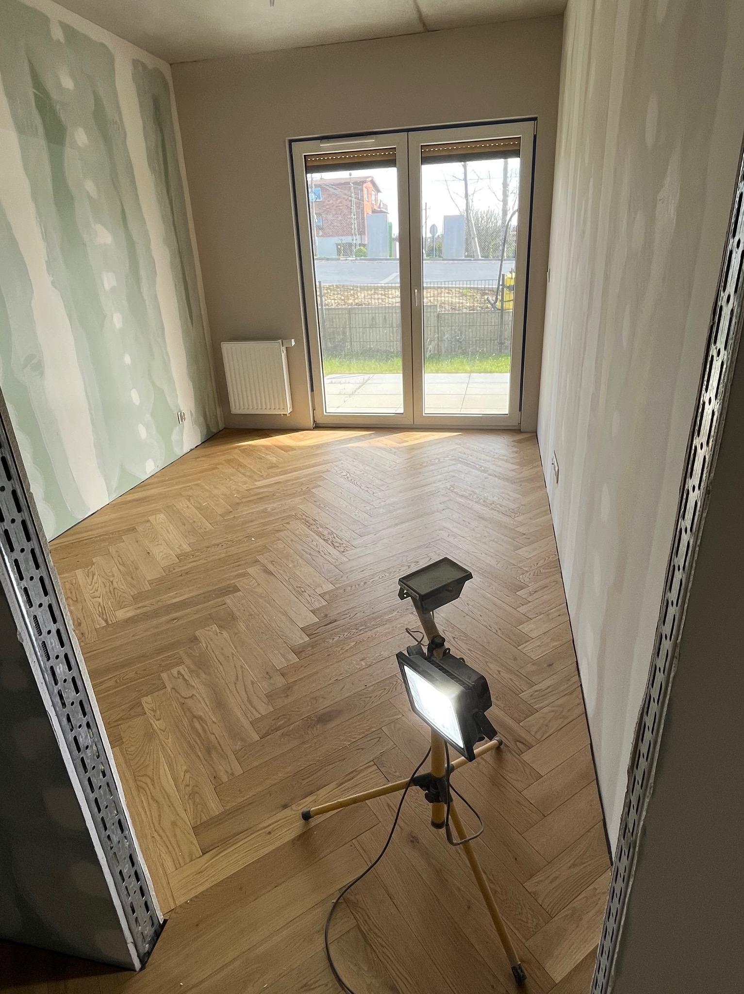 Jasny parkiet ułożony w jodełkę w pokoju w trakcie wykańczania. Ściany częściowo przygotowane do malowania, widoczna lampa budowlana.