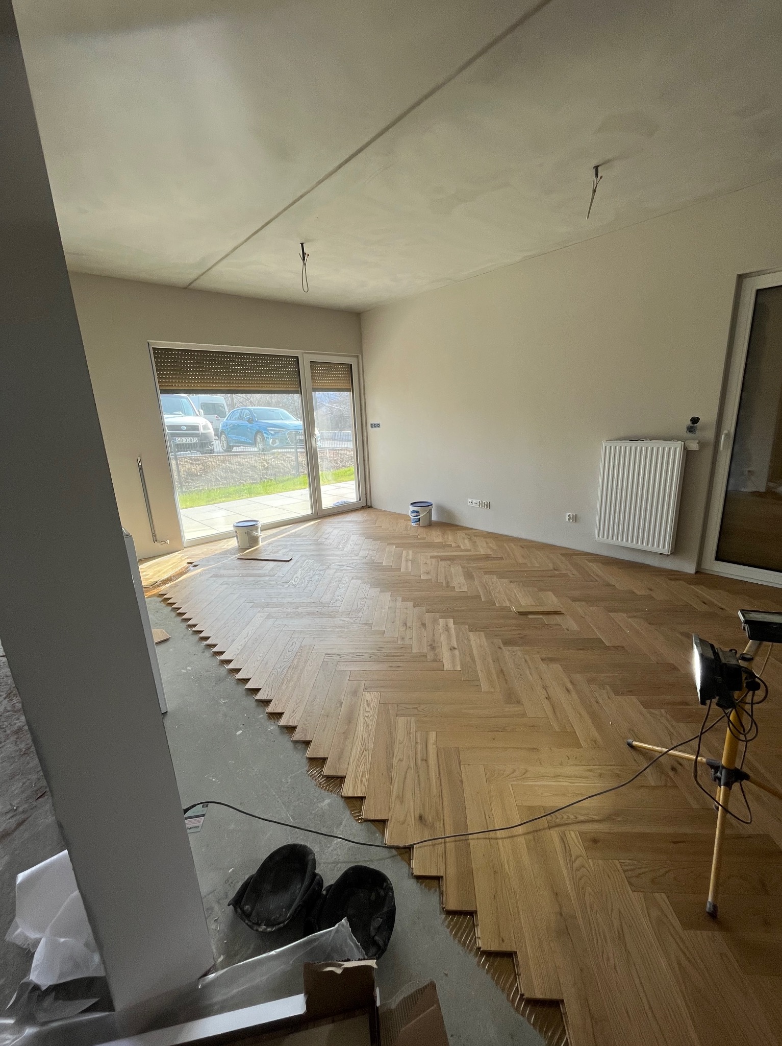 Układanie parkietu w jodełkę w Bielsko-Białej. Częściowo ułożona podłoga z drewnianych klepek, widoczne narzędzia i lampa oświetleniowa. Jasne wnętrze z oknem.