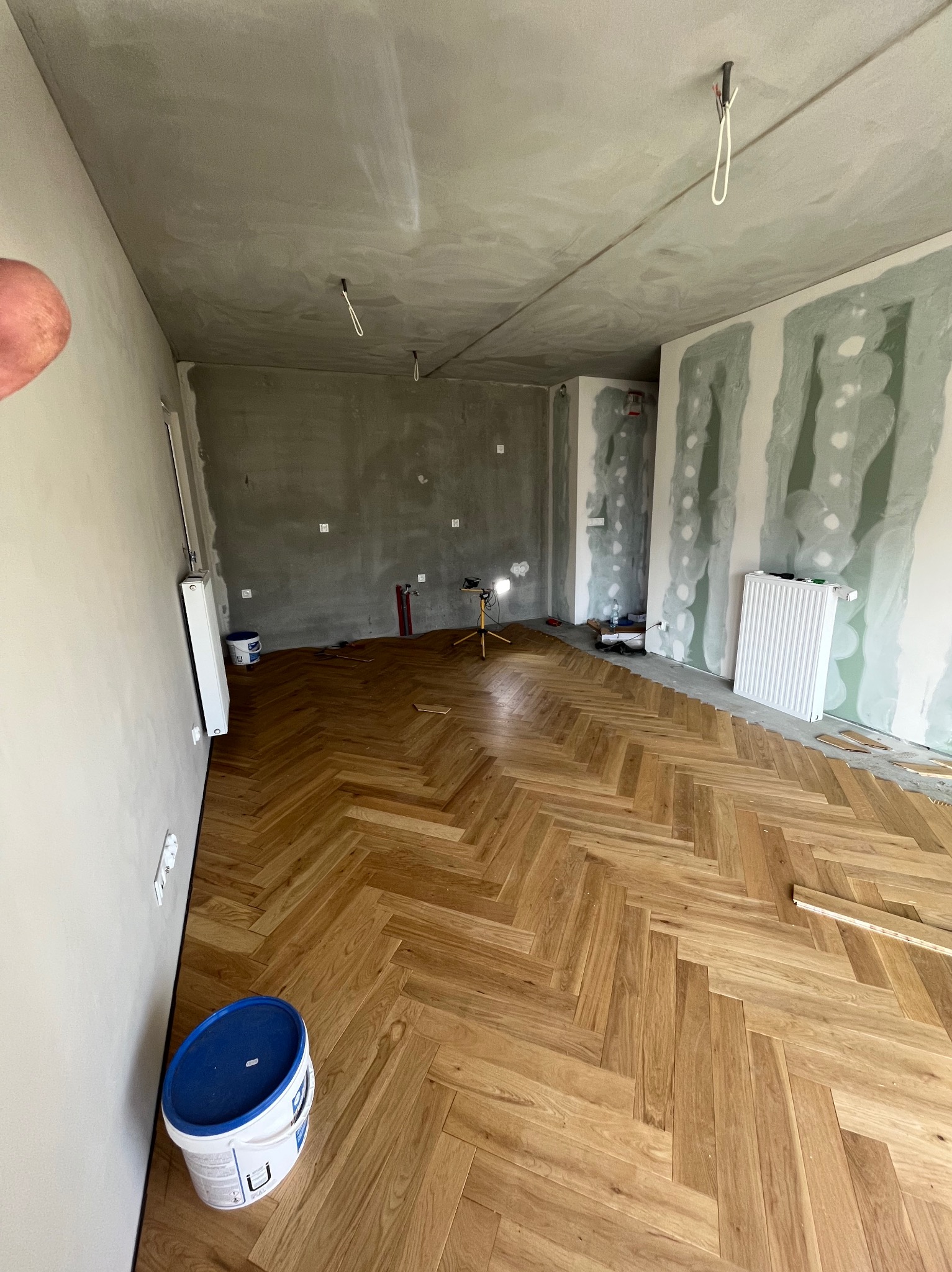 Układanie parkietu w jodełkę w surowym wnętrzu. Widoczne przygotowane ściany, grzejnik i wiadro farby. Oświetlenie budowlane w tle.