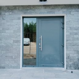 Przemysław Suchecki - Elewacja domu z szarej cegły z nowoczesnymi, stalowymi drzwiami wejściowymi i dużym oknem, odbijającym las i biały samochód dostawczy.