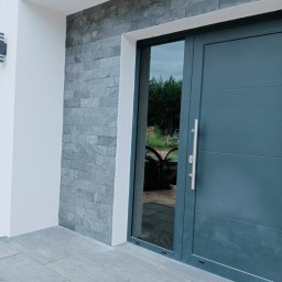 Przemysław Suchecki - Eleganckie wejście do domu w Świebodzinie z szarą, nowoczesną stolarką drzwiową i kamienną okładziną ściany. Widoczny fragment chodnika z szarej kostki.