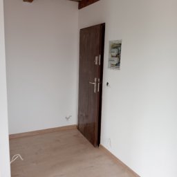 Przemysław Suchecki - Wnętrze po remoncie: drewniana podłoga, białe ściany z brązowymi listwami, ciemne drzwi i skrzynka elektryczna. Widoczne drewniane belki stropowe.