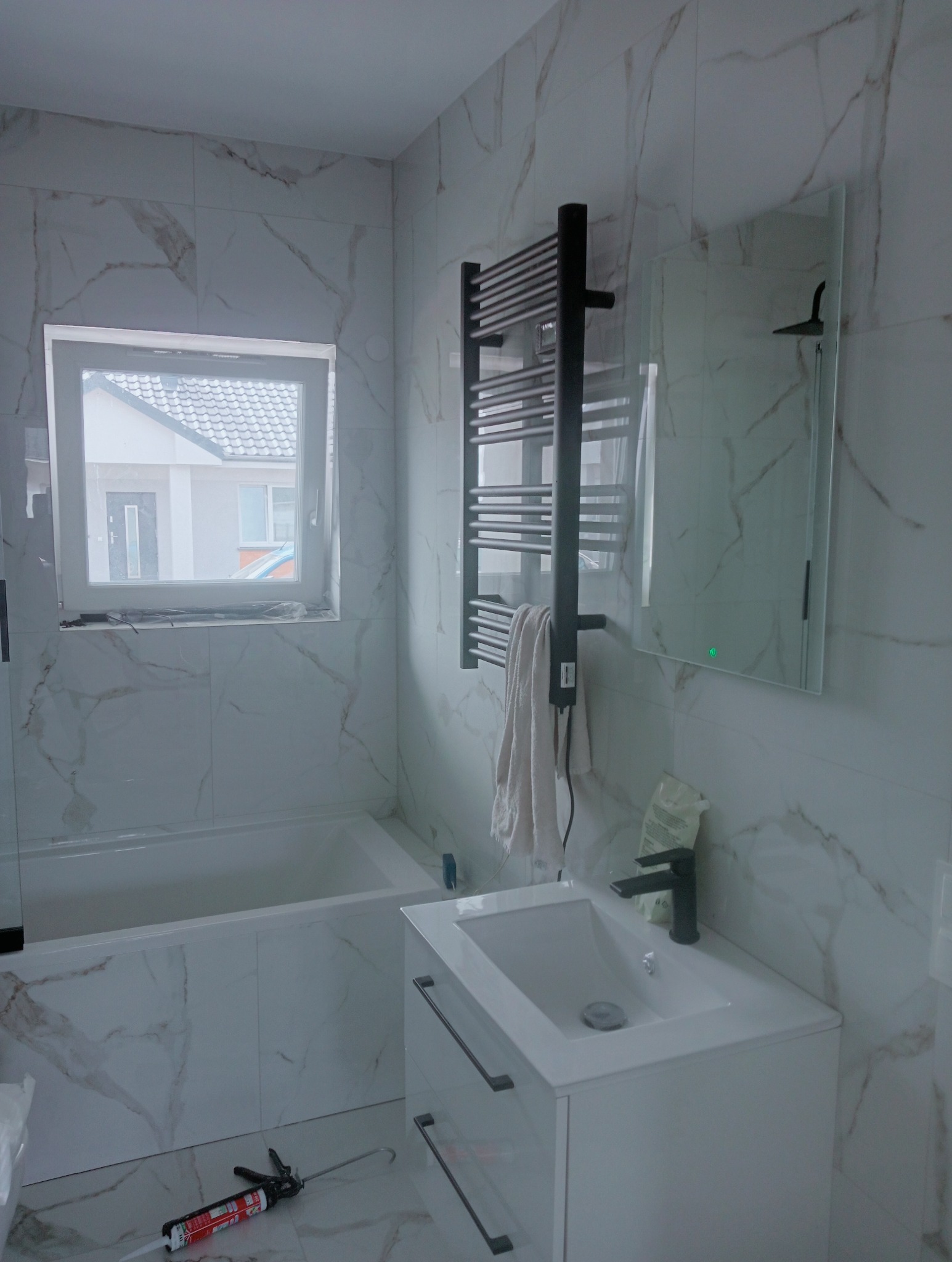 Wykończona łazienka z wanną, umywalką z szafką i grzejnikiem drabinkowym. Białe płytki imitujące marmur na ścianach i podłodze. Widoczne narzędzia.