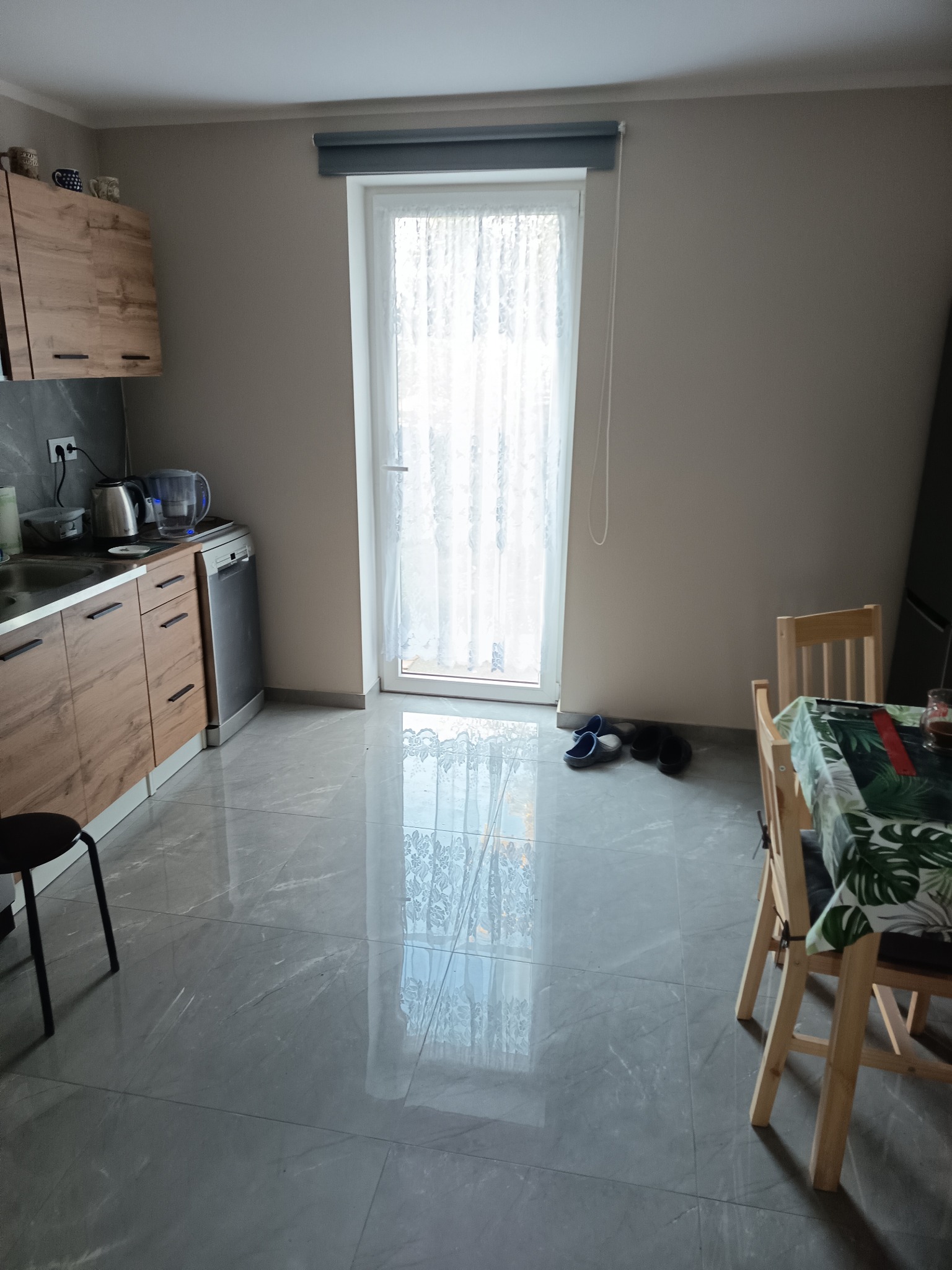 Jasna kuchnia z szarymi płytkami podłogowymi, drewnianymi szafkami i oknem z firanką. Widoczne krzesła i stół z zielonym obrusem. Minimalistyczny wystrój.