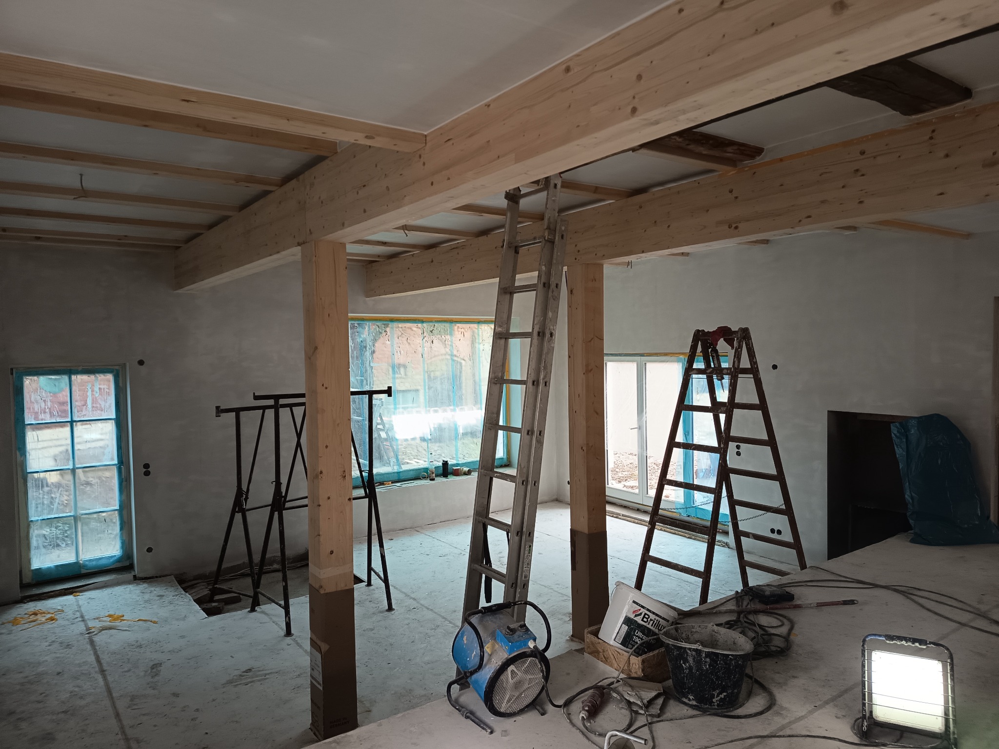 Wnętrze w trakcie remontu: drewniane belki stropowe, drabiny, narzędzia malarskie, ściany po gładziowaniu, okna zabezpieczone folią. Widoczne przygotowania do dalszych prac wykończeniowych.