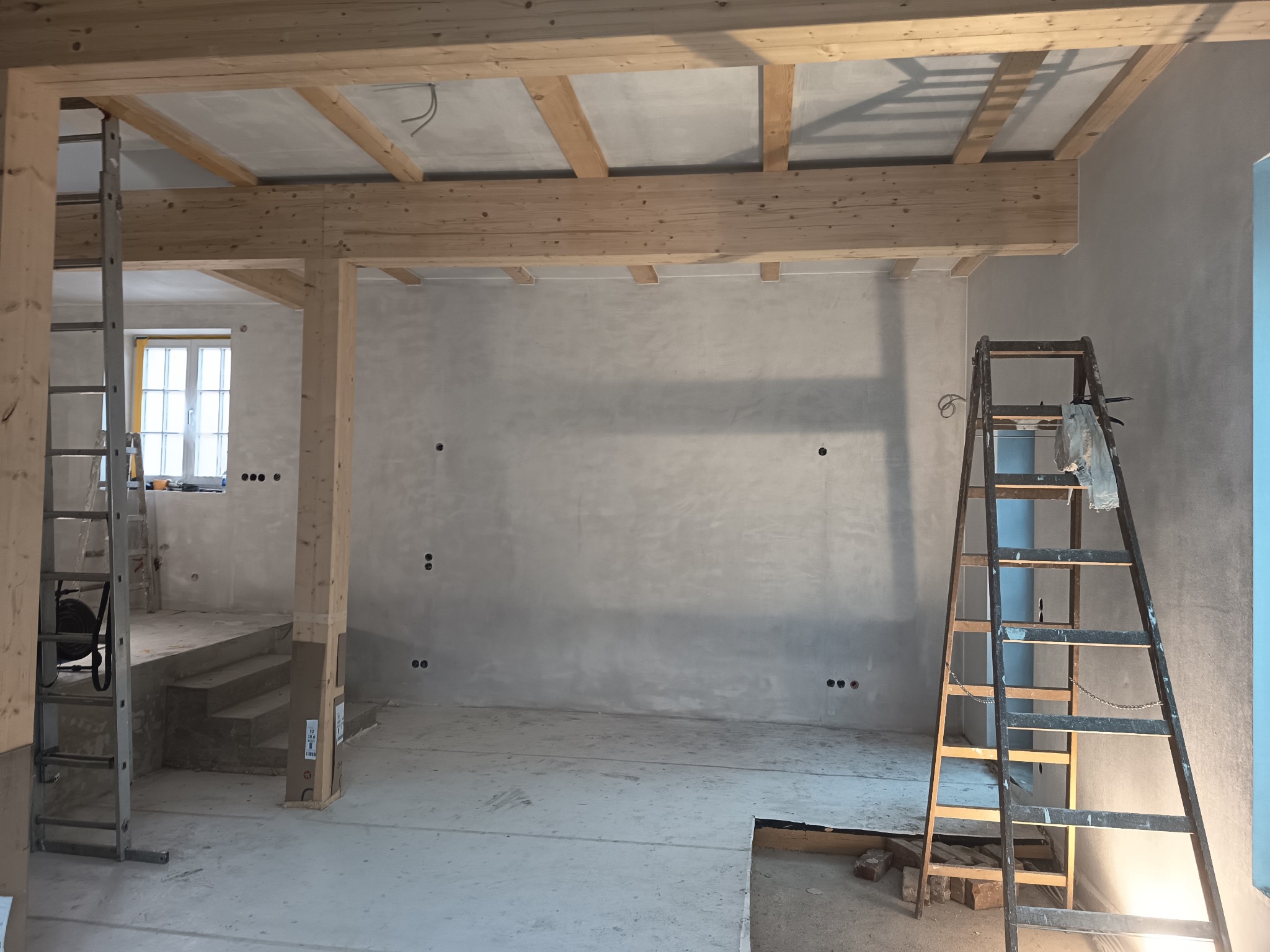 Surowy stan wnętrza z drewnianą konstrukcją stropu, gładzią na ścianach i dwiema drabinami. Widoczne przygotowania do dalszych prac wykończeniowych.