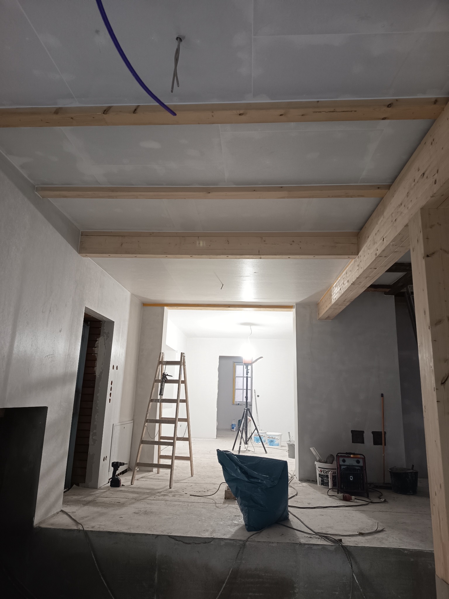 Wnętrze w trakcie remontu: sufit z płyt kartonowo-gipsowych, drewniane belki, ściany po gładziowaniu, drabina i narzędzia. Widok na kolejne pomieszczenie z oknem.