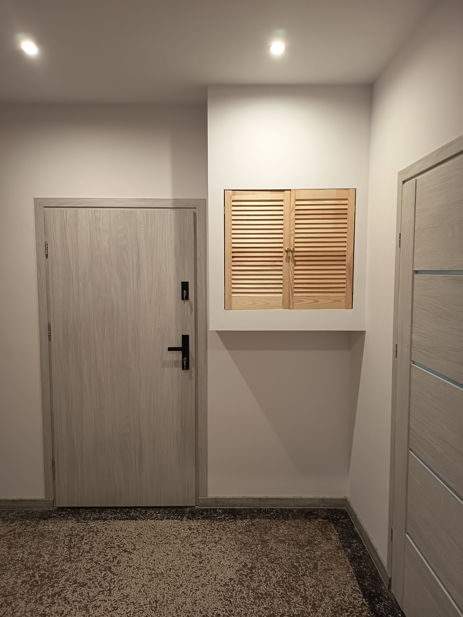 Jasne drzwi wejściowe z czarną klamką i drewniane żaluzje wnękowe w białej ścianie, widoczne w korytarzu z wzorzystą podłogą.