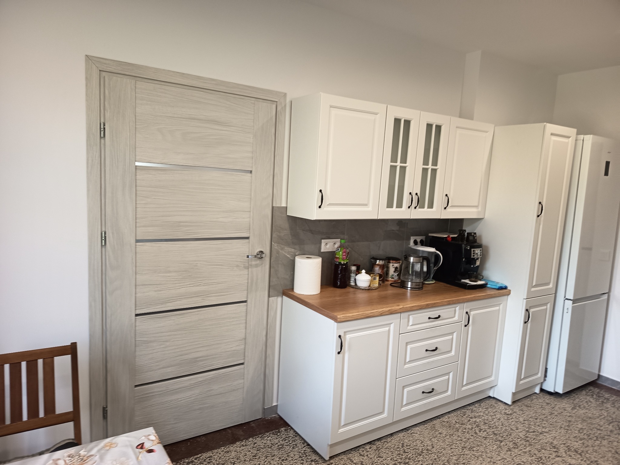 Jasna kuchnia z drewnianym blatem i białymi szafkami, widok na drzwi z poziomymi panelami i fragment brązowego krzesła w rogu.