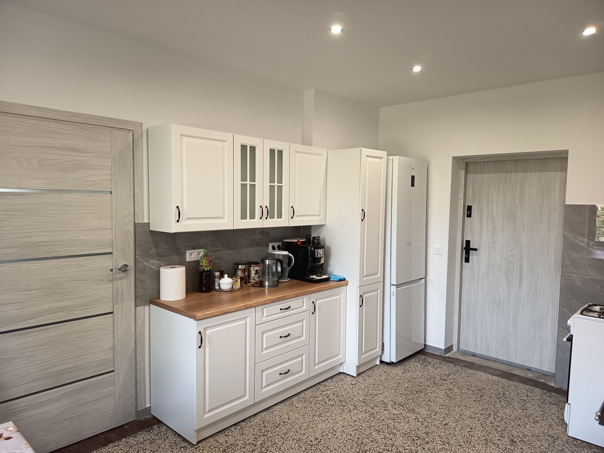 Jasna kuchnia z drewnianym blatem, białymi szafkami i sprzętem AGD. Widoczne drzwi z szarą okleiną i mozaikowa podłoga. Minimalistyczny styl.