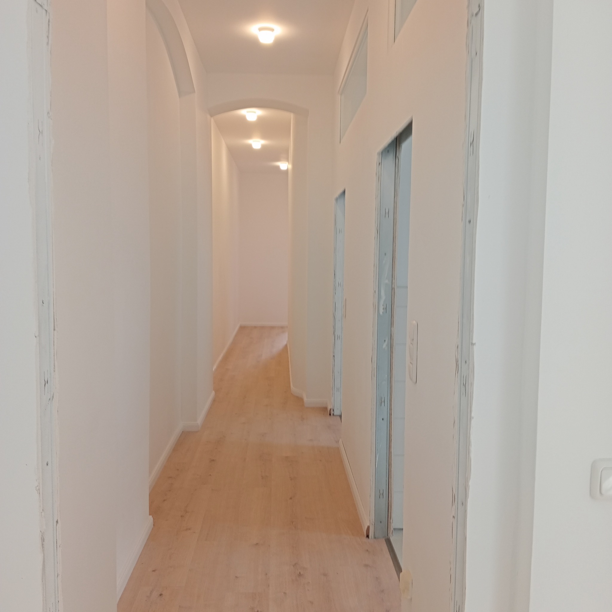 Długi, jasny korytarz po remoncie z drewnianą podłogą i świeżo pomalowanymi ścianami. Widoczne łuki i wnęki w ścianach. Oświetlenie sufitowe.