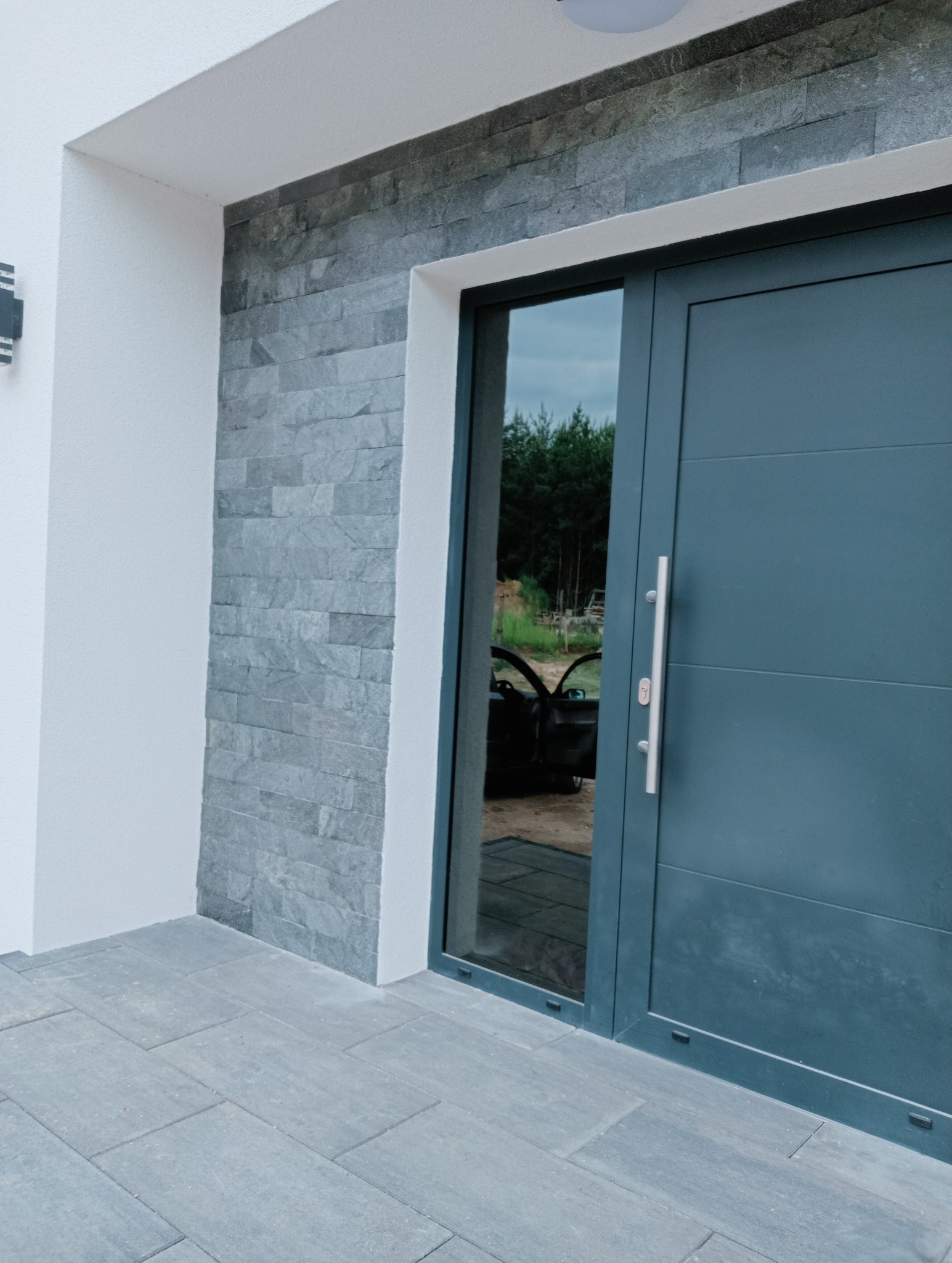Eleganckie wejście do domu w Świebodzinie z szarą, nowoczesną stolarką drzwiową i kamienną okładziną ściany. Widoczny fragment chodnika z szarej kostki.