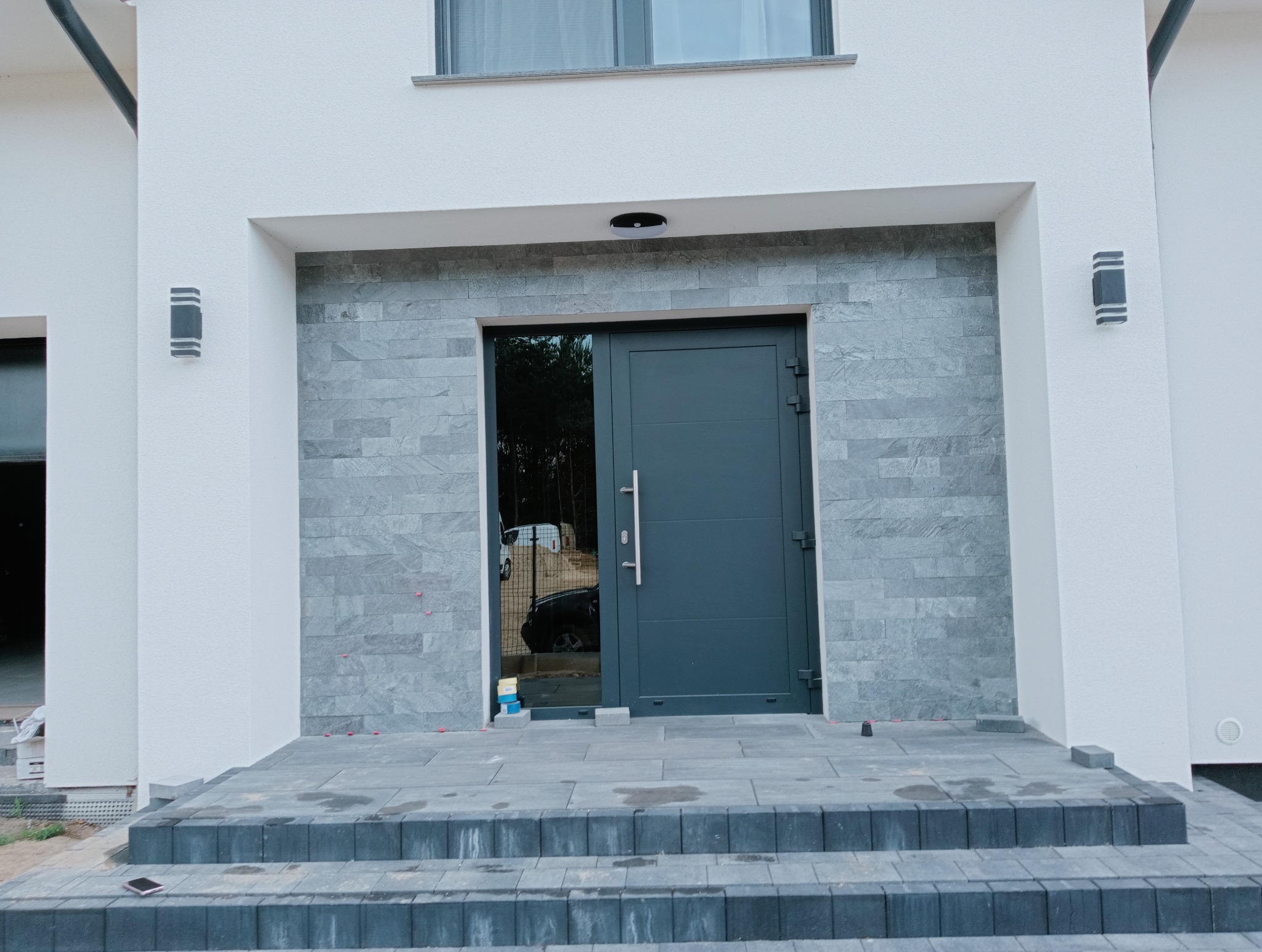 Nowoczesne wejście do domu z szarą elewacją z kamienia, ciemnoszare drzwi z długim uchwytem i schody z kostki brukowej. Widoczne lampy zewnętrzne.