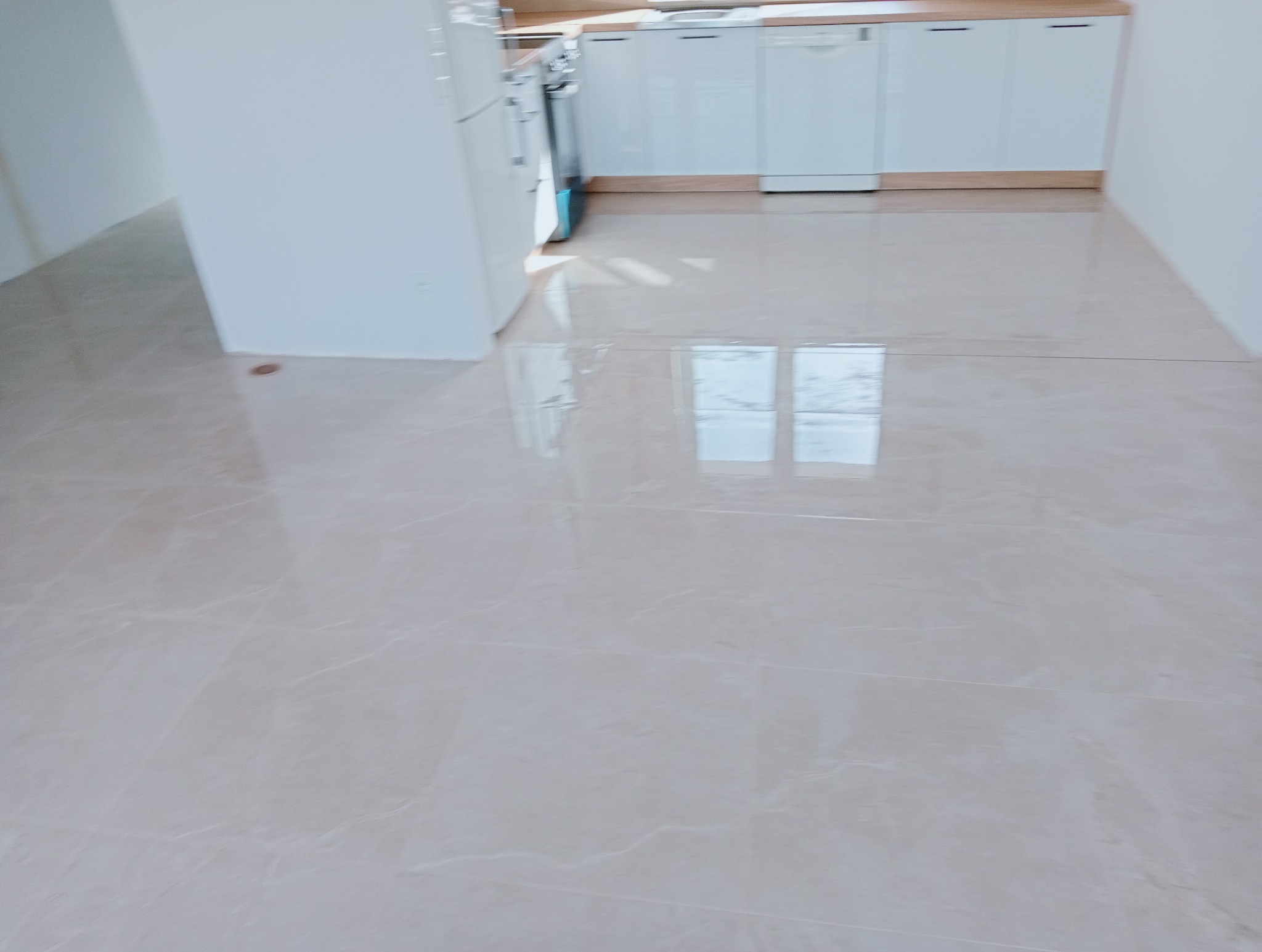 Jasne, błyszczące płytki podłogowe w kuchni z białymi szafkami i drewnianym blatem. Widoczne odbicia światła na podłodze. Minimalistyczny styl.