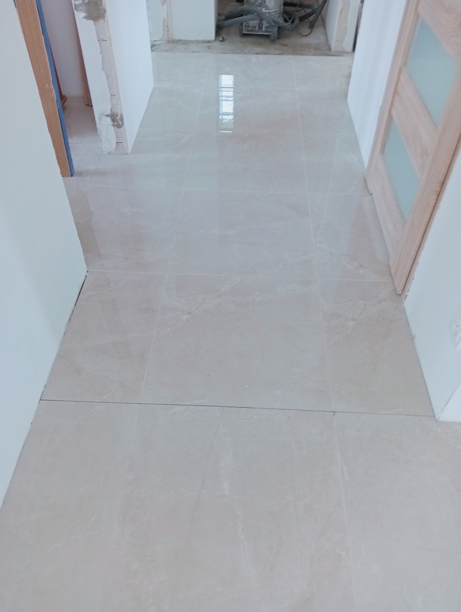 Jasne, błyszczące płytki podłogowe w korytarzu, świeżo po ułożeniu. Widoczne drzwi z drewnianą ramą i fragmenty ścian w trakcie wykończenia. W tle sprzęt budowlany.