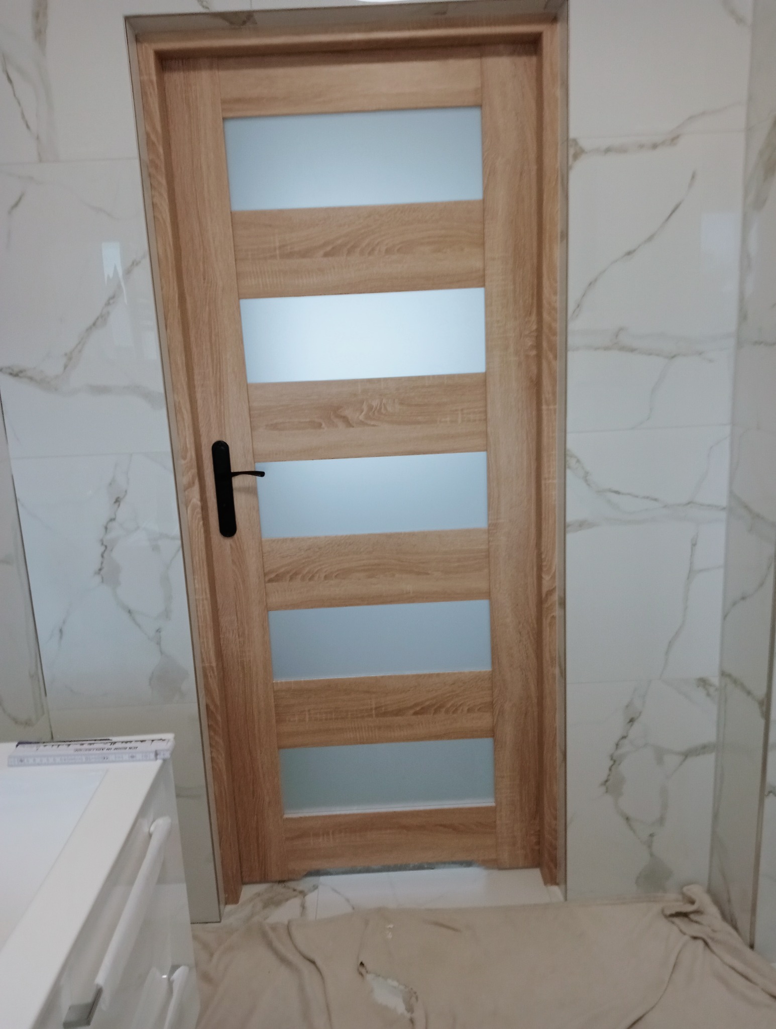 Zamknięte drzwi wewnętrzne z drewnianą ramą i czterema poziomymi, matowymi szybami, zamontowane w łazience z marmurowymi płytkami. Widoczna czarna klamka.