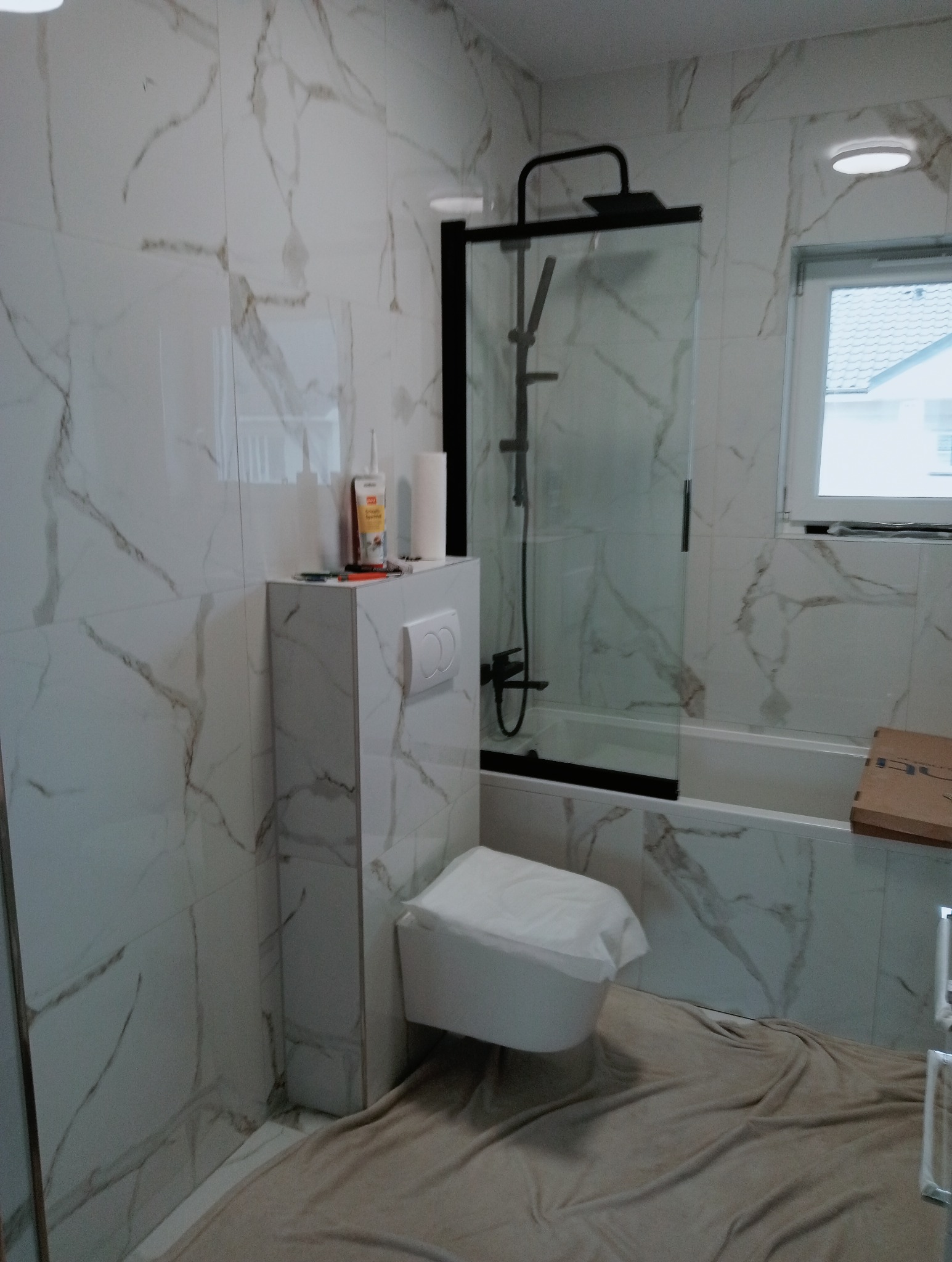 Wykończona łazienka z marmurowymi płytkami, czarną armaturą i podwieszaną toaletą. Podłoga zabezpieczona folią malarską. Widoczne akcesoria montażowe.