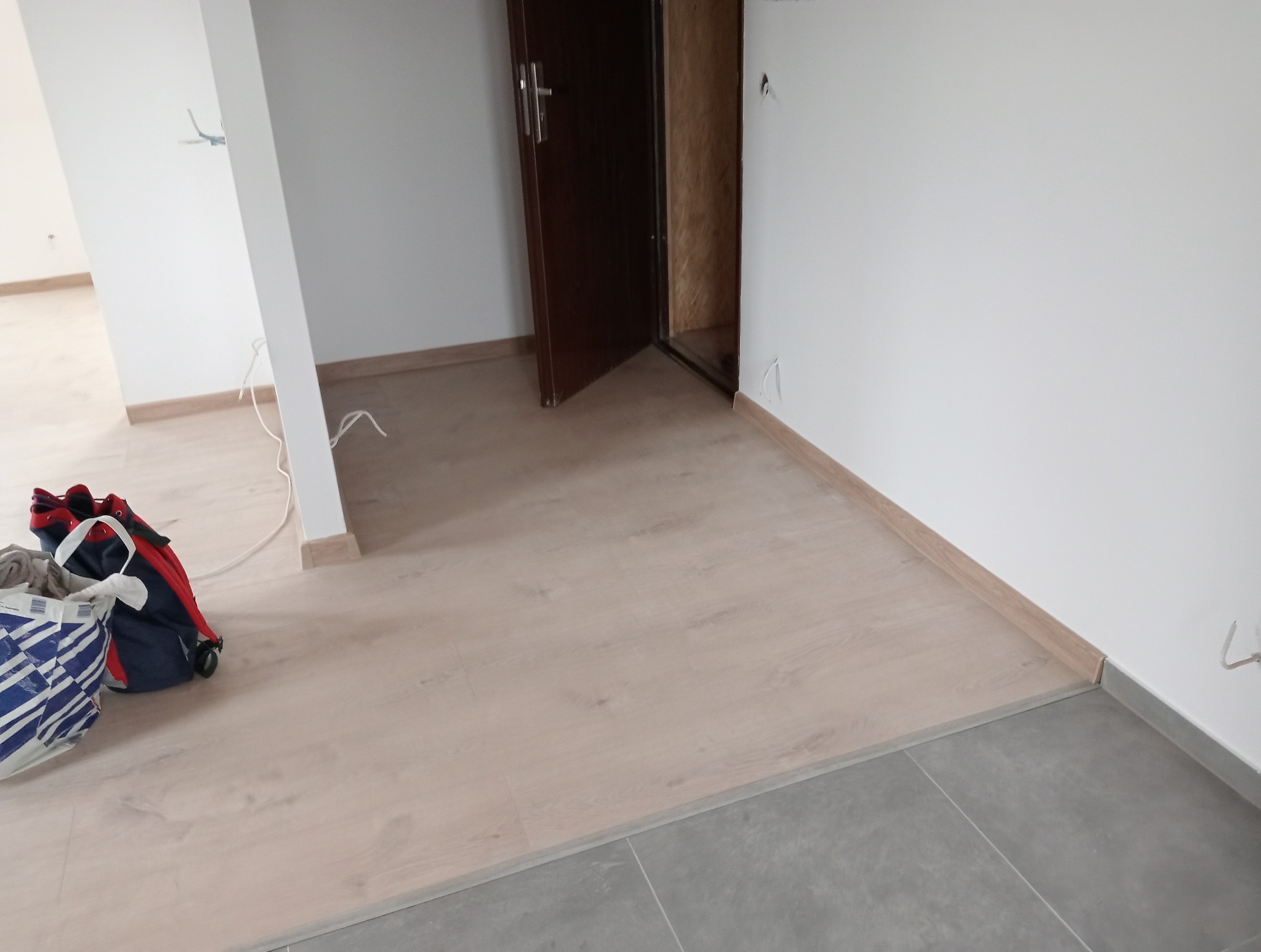 Jasne panele podłogowe przechodzą w szare płytki, widoczna torba robocza, białe ściany i brązowe drzwi. Fragment wykończonego wnętrza po remoncie.