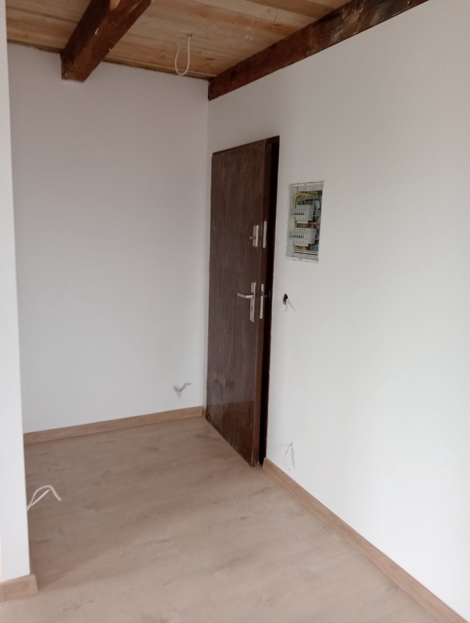 Wnętrze po remoncie: drewniana podłoga, białe ściany z brązowymi listwami, ciemne drzwi i skrzynka elektryczna. Widoczne drewniane belki stropowe.
