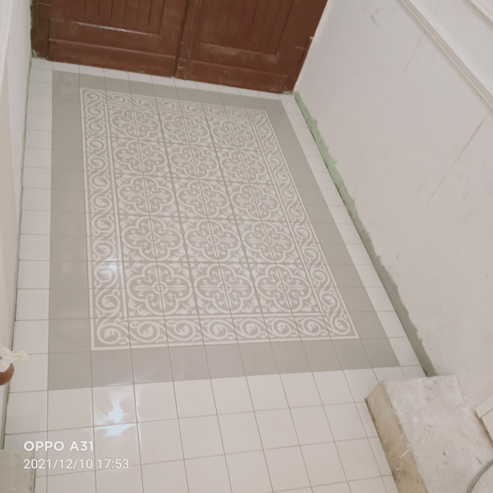 Ułożona podłoga z płytek w łazience z dekoracyjnym wzorem w centralnej części. Widoczny fragment drzwi i ściany w trakcie wykończenia.